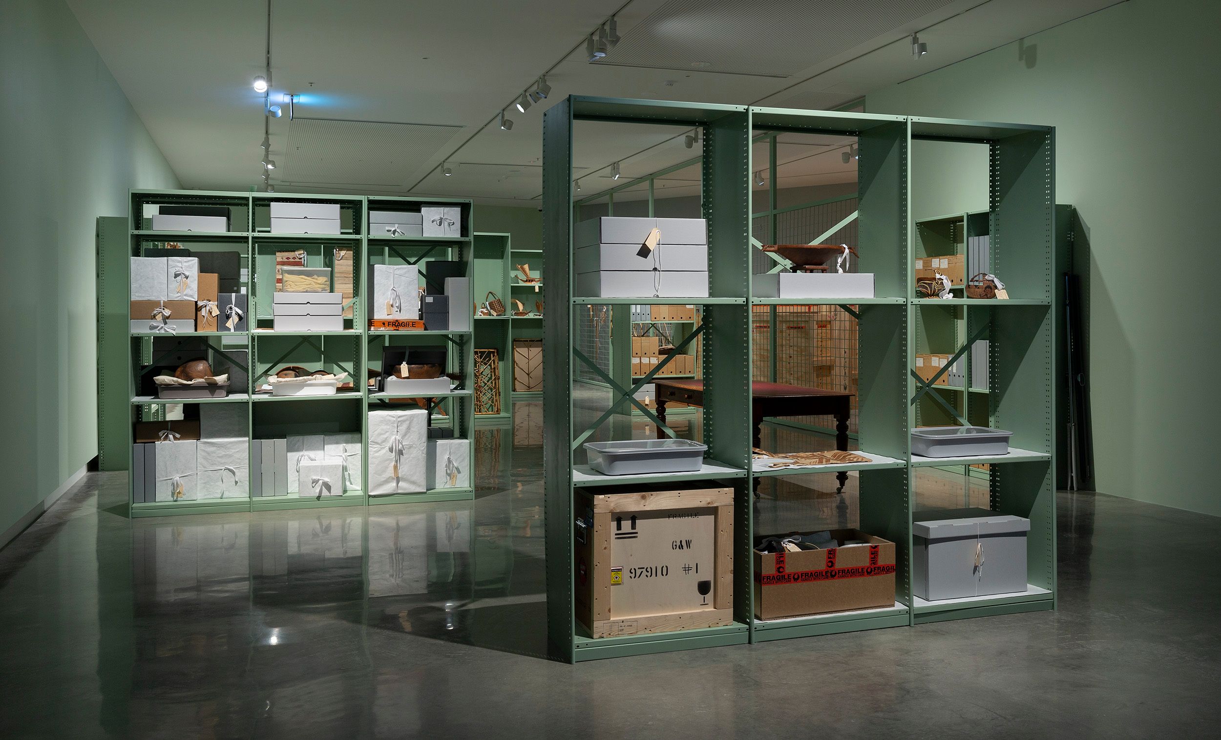 A modern gallery space with green metal shelving units displaying various boxes, crates, and wrapped objects, creating an organized, storage-like art installation. The floor is shiny and the walls are pale green.