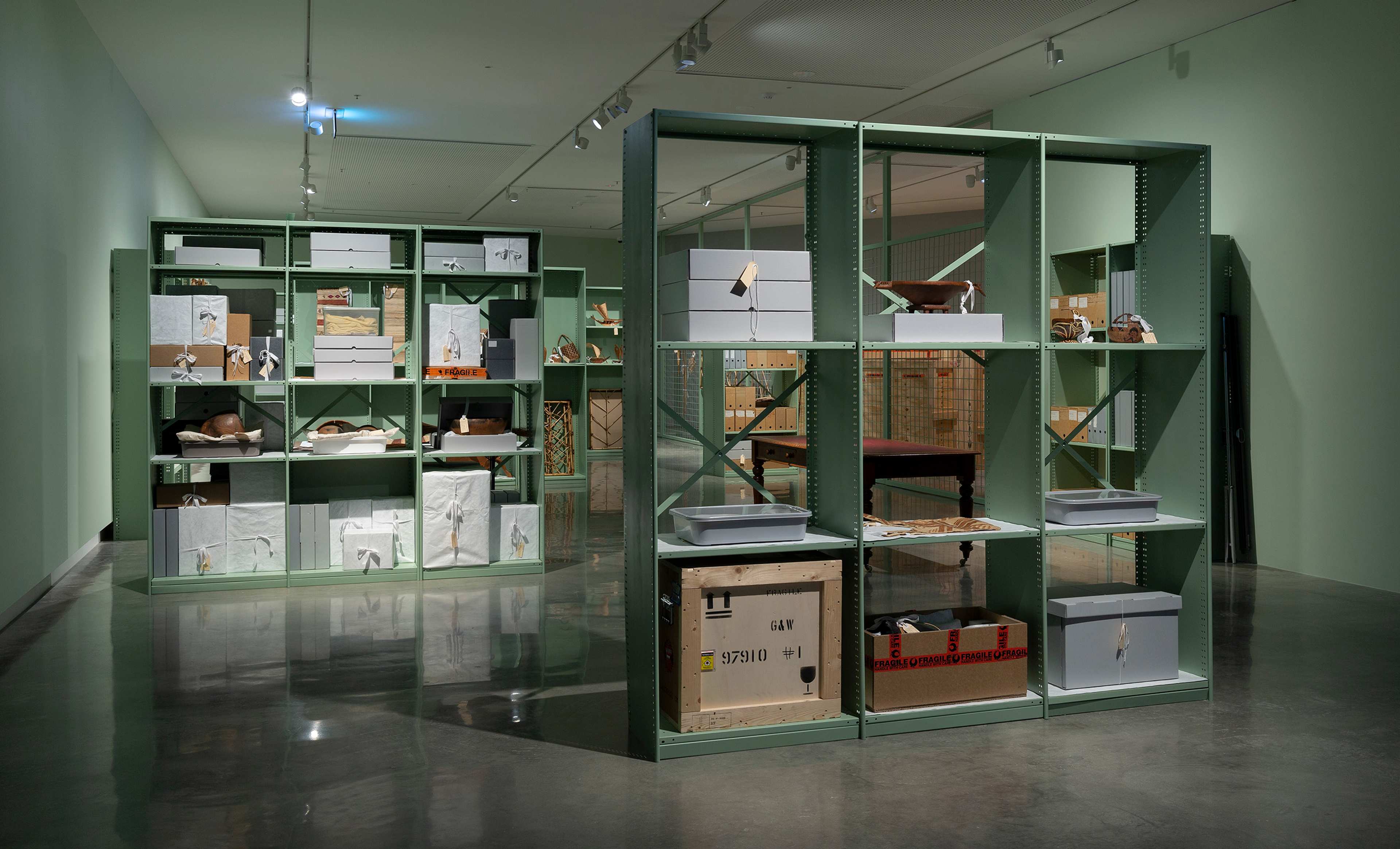 A modern gallery space with green metal shelving units displaying various boxes, crates, and wrapped objects, creating an organized, storage-like art installation. The floor is shiny and the walls are pale green.
