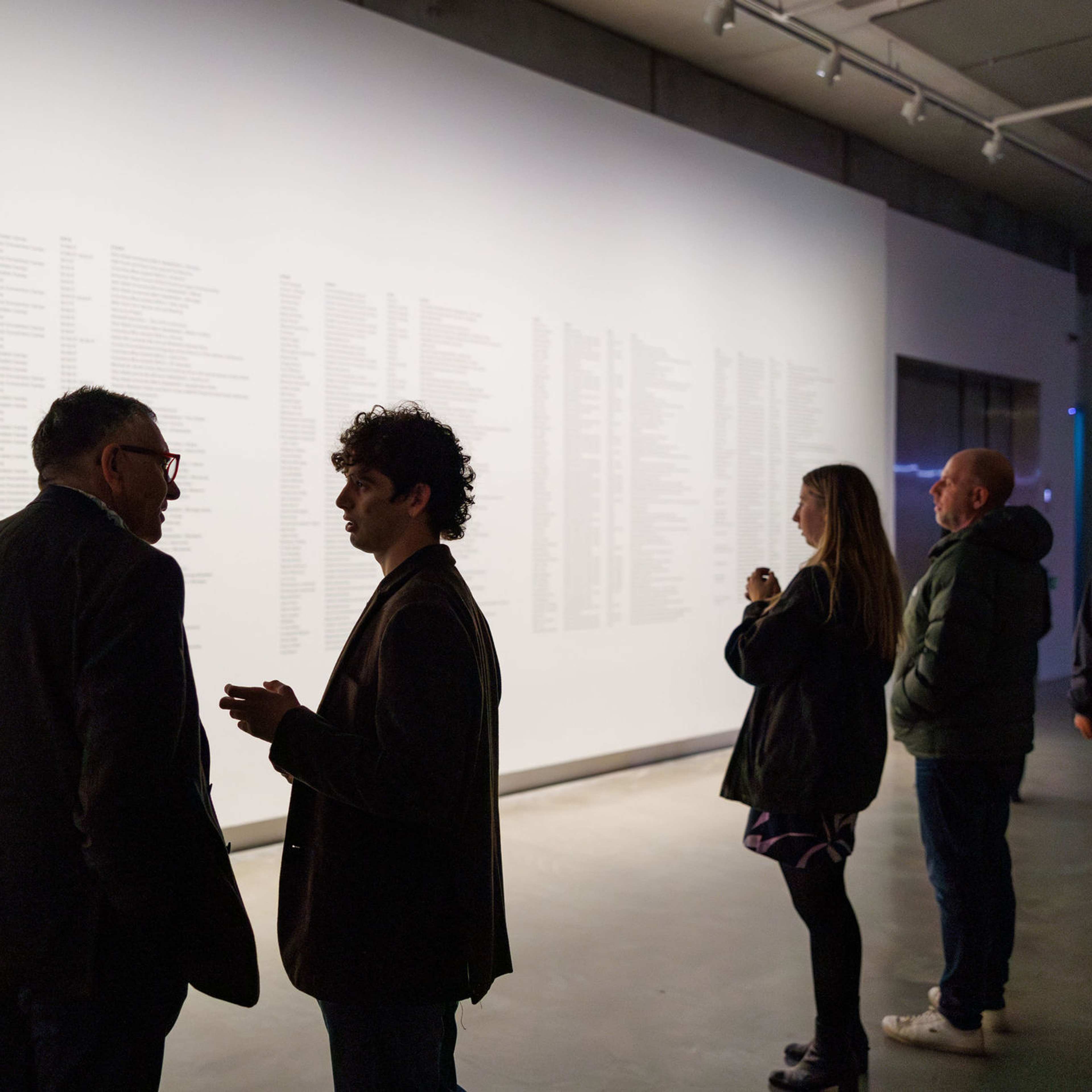 People stand in a dimly lit gallery looking at a large, illuminated wall covered with columns of text. Some individuals talk while others observe the display quietly.