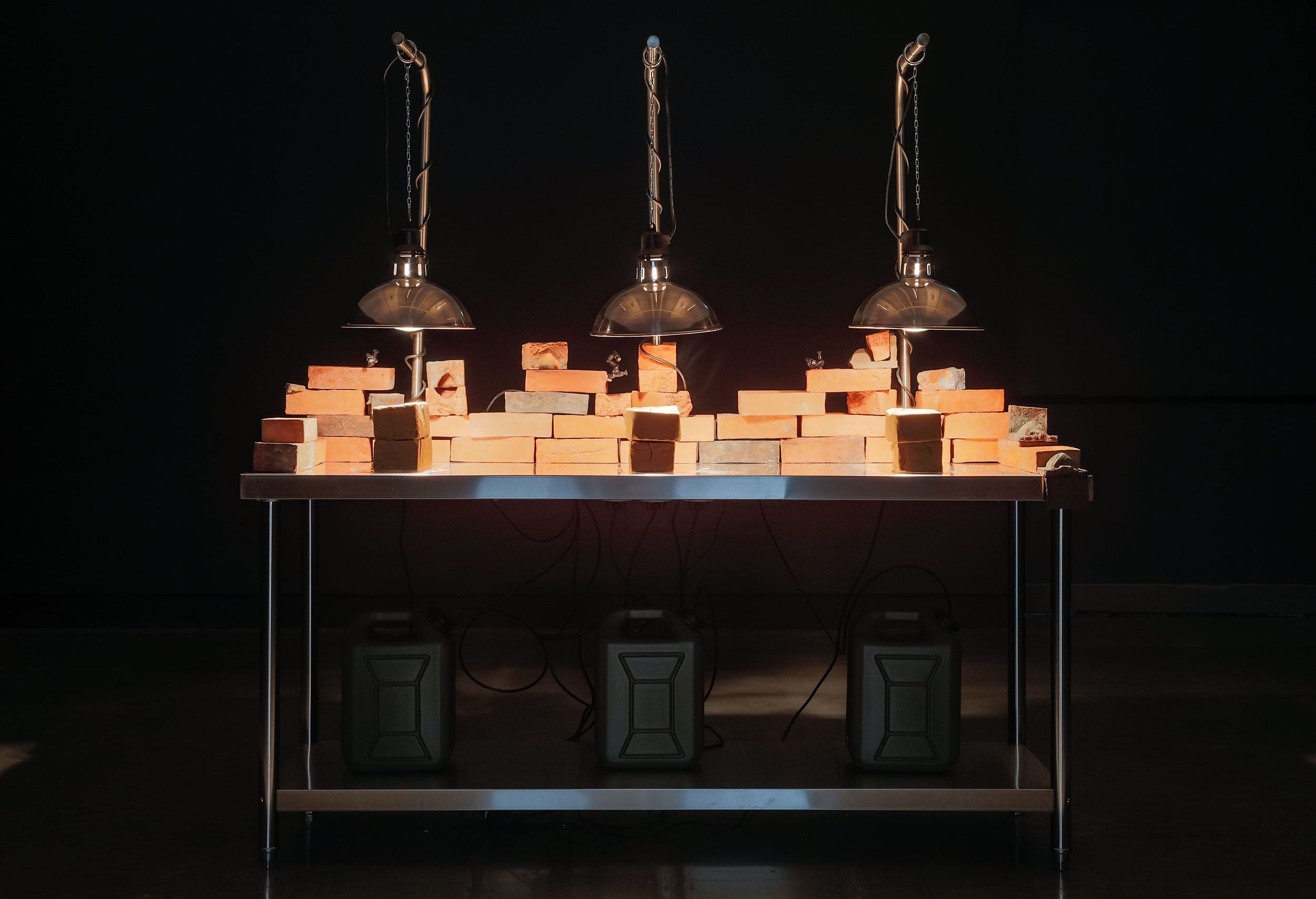 A metal table holds three rows of bricks illuminated by three hanging heat lamps. Under the table are three rectangular containers with handles, and electrical wires are visible in the dimly lit setting.