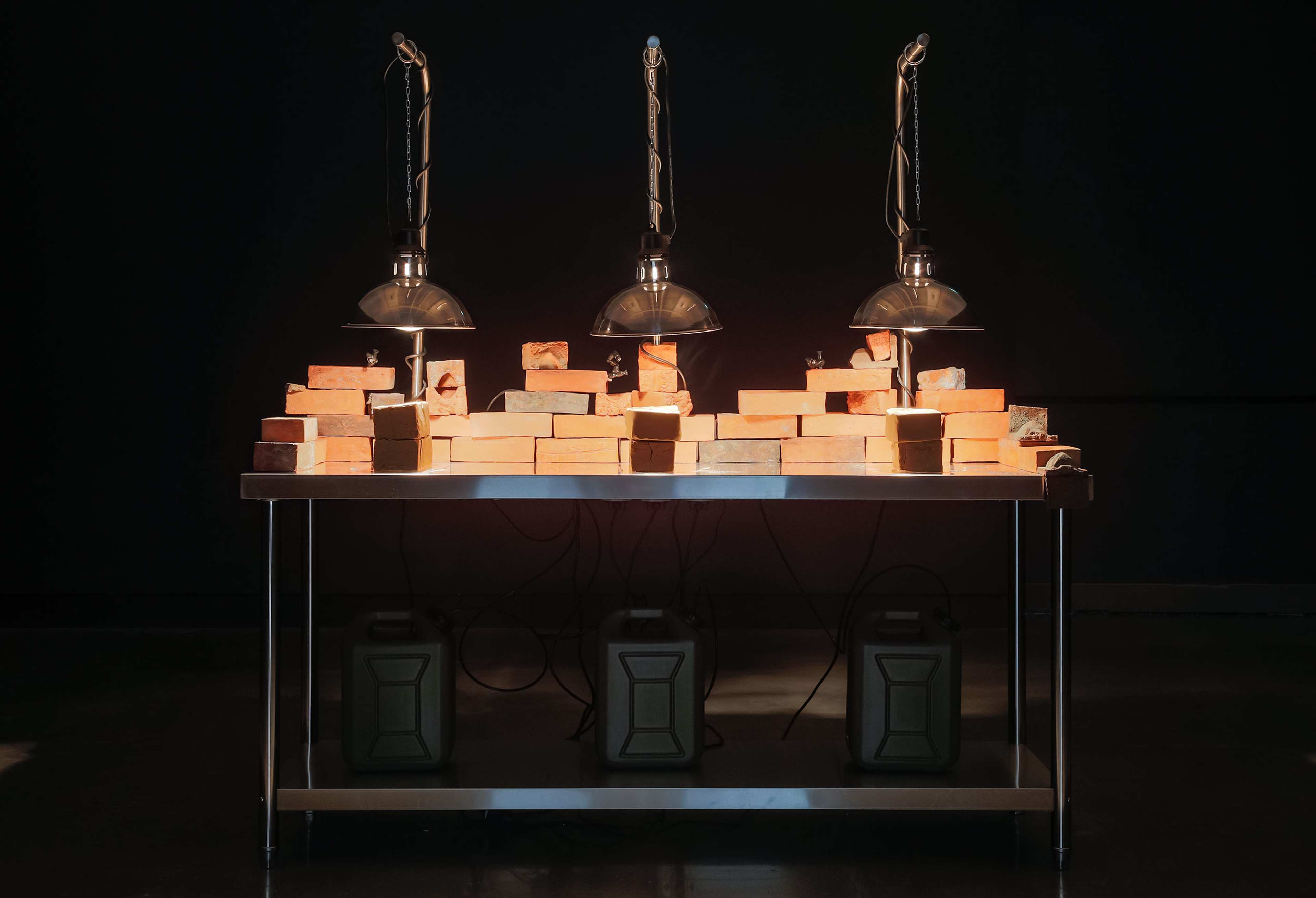 A metal table holds three rows of bricks illuminated by three hanging heat lamps. Under the table are three rectangular containers with handles, and electrical wires are visible in the dimly lit setting.