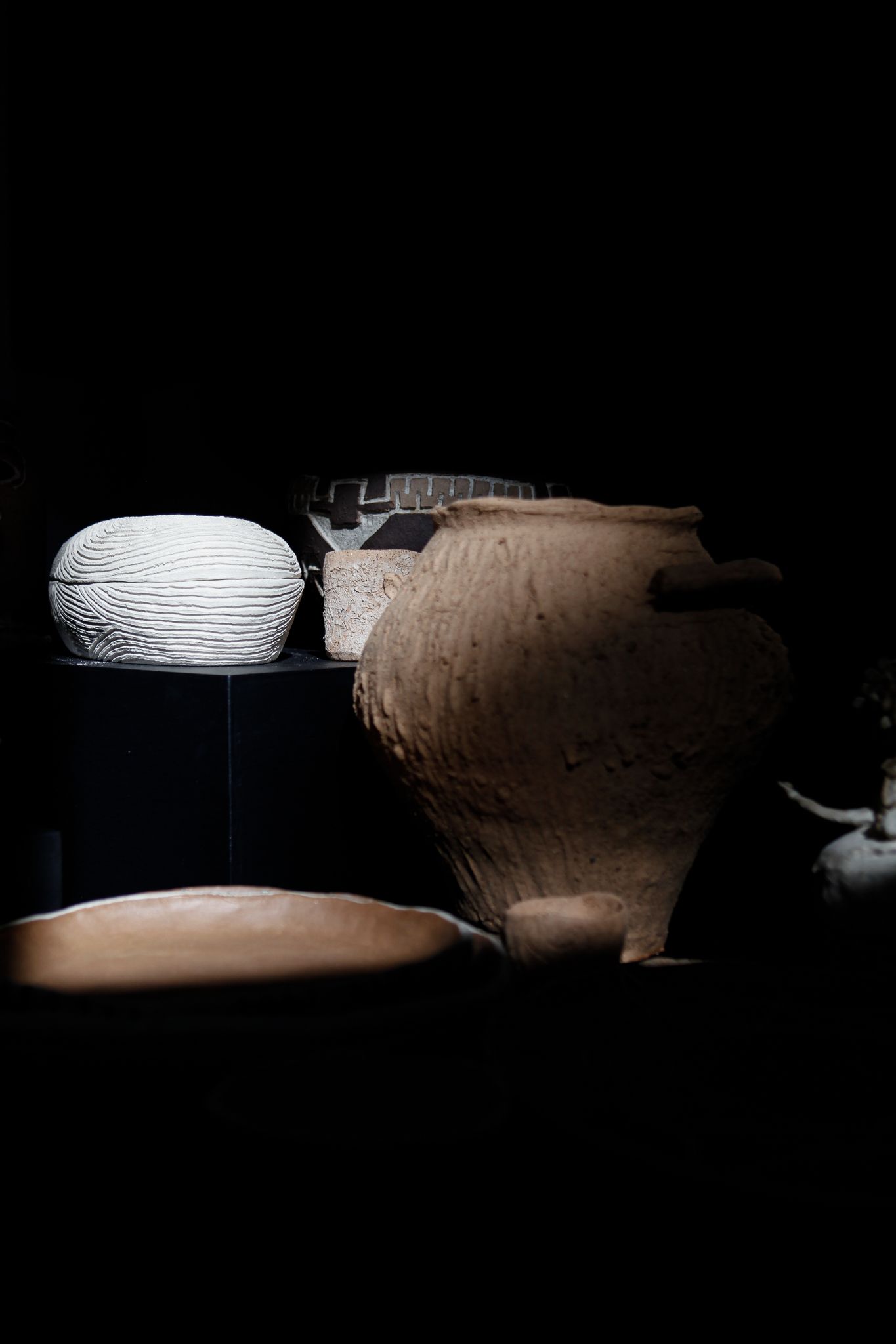 A group of ancient pottery vessels, including a textured clay jar, a white ribbed bowl, and other earthenware, displayed against a dark background with dramatic lighting highlighting their surfaces.