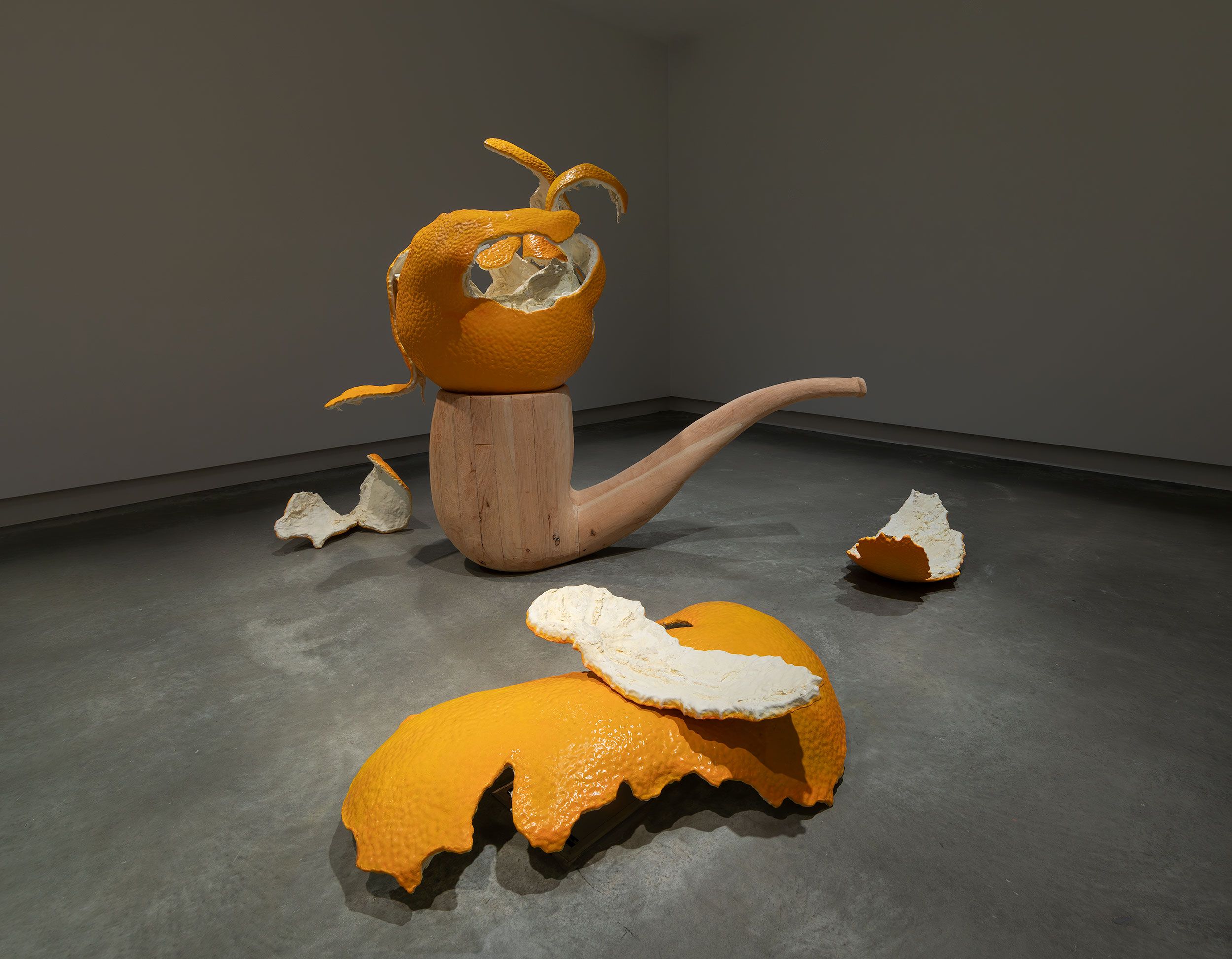 A large wooden sculpture shaped like a smoking pipe is topped with a hollowed orange peel, with peeled orange pieces scattered on the smooth gray floor in a minimalist gallery setting.