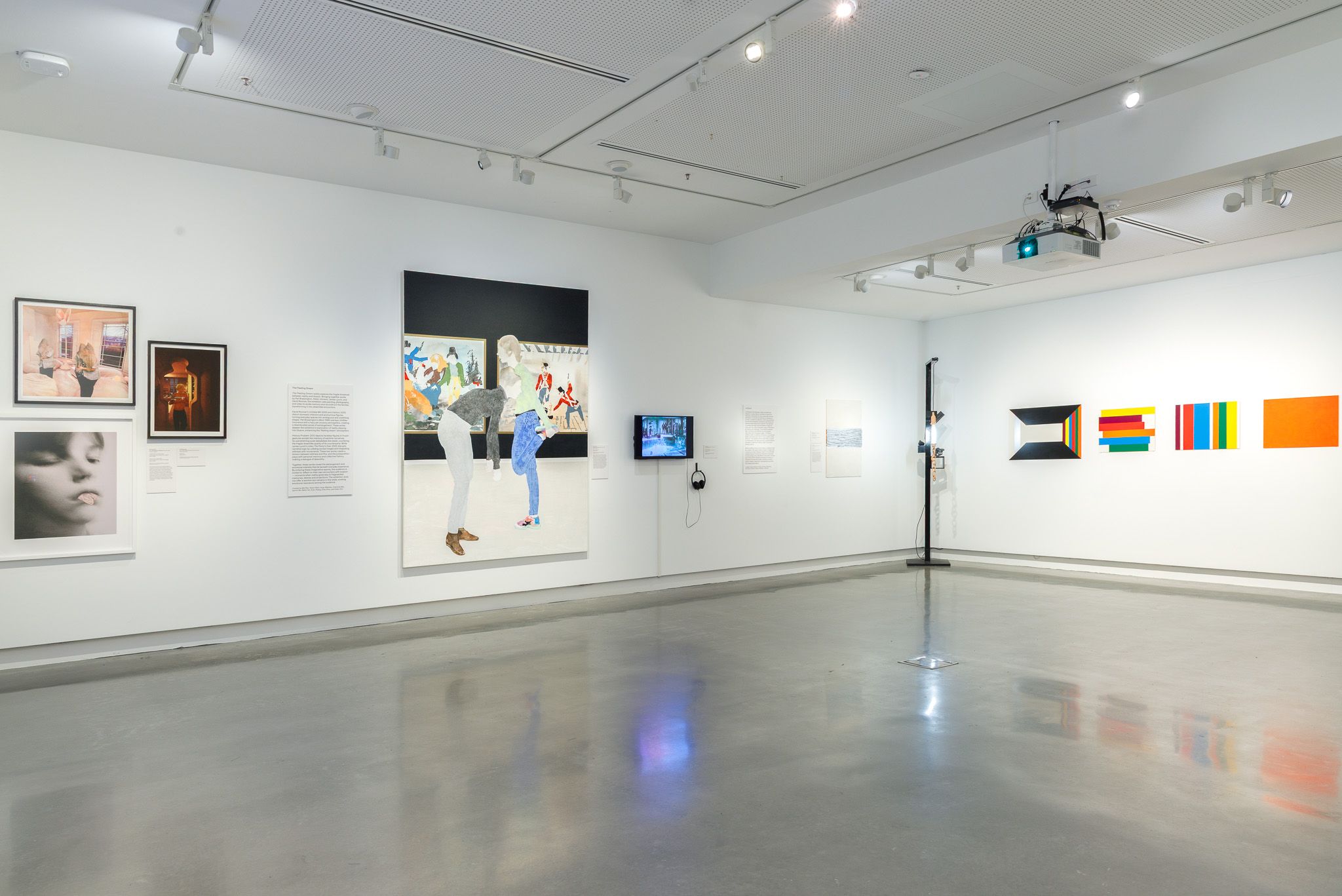 A modern art gallery with white walls displays paintings, photographs, and a video screen. The floor is polished concrete, and the space is brightly lit with overhead lights. No people are present.