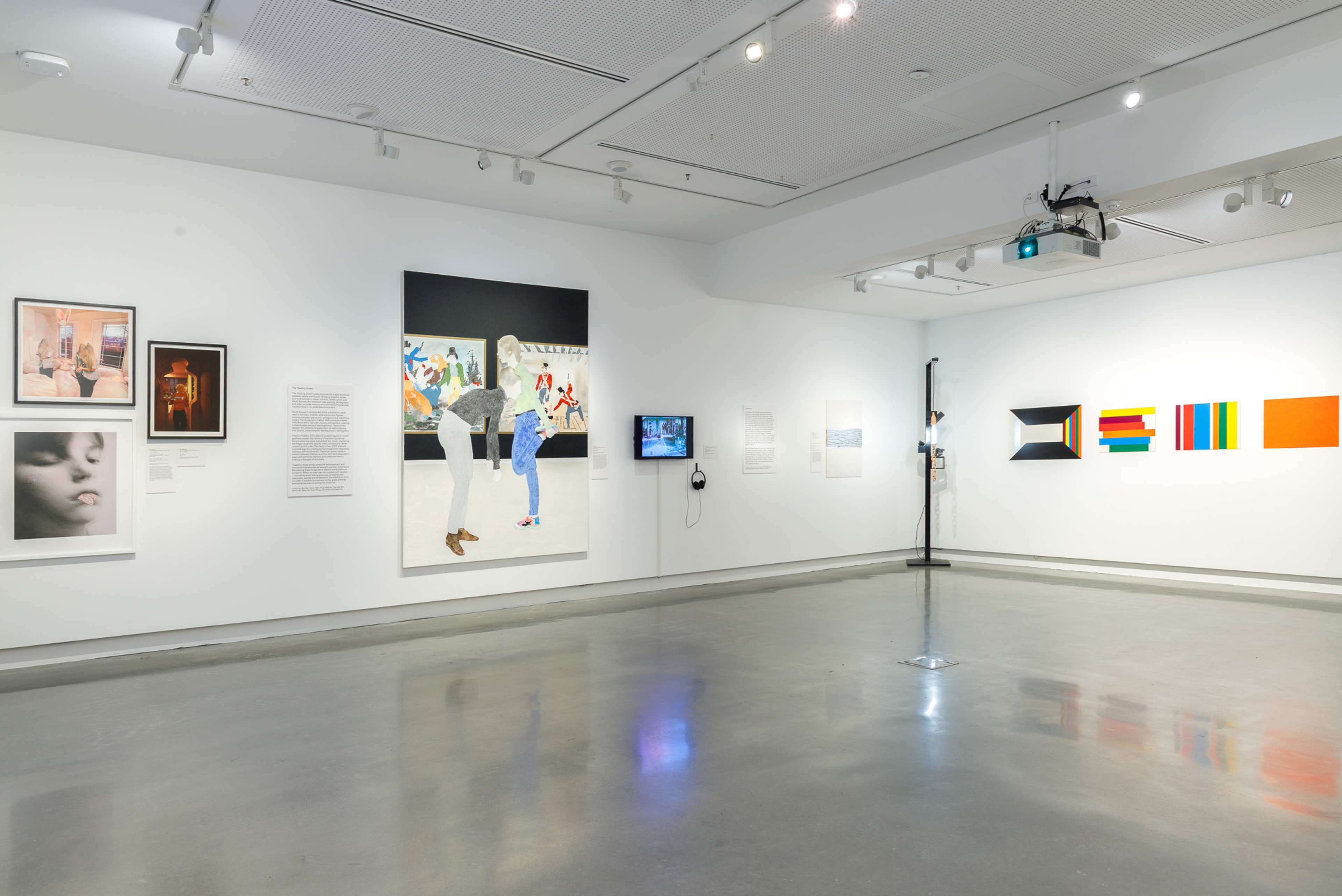 A modern art gallery with white walls displays paintings, photographs, and a video screen. The floor is polished concrete, and the space is brightly lit with overhead lights. No people are present.