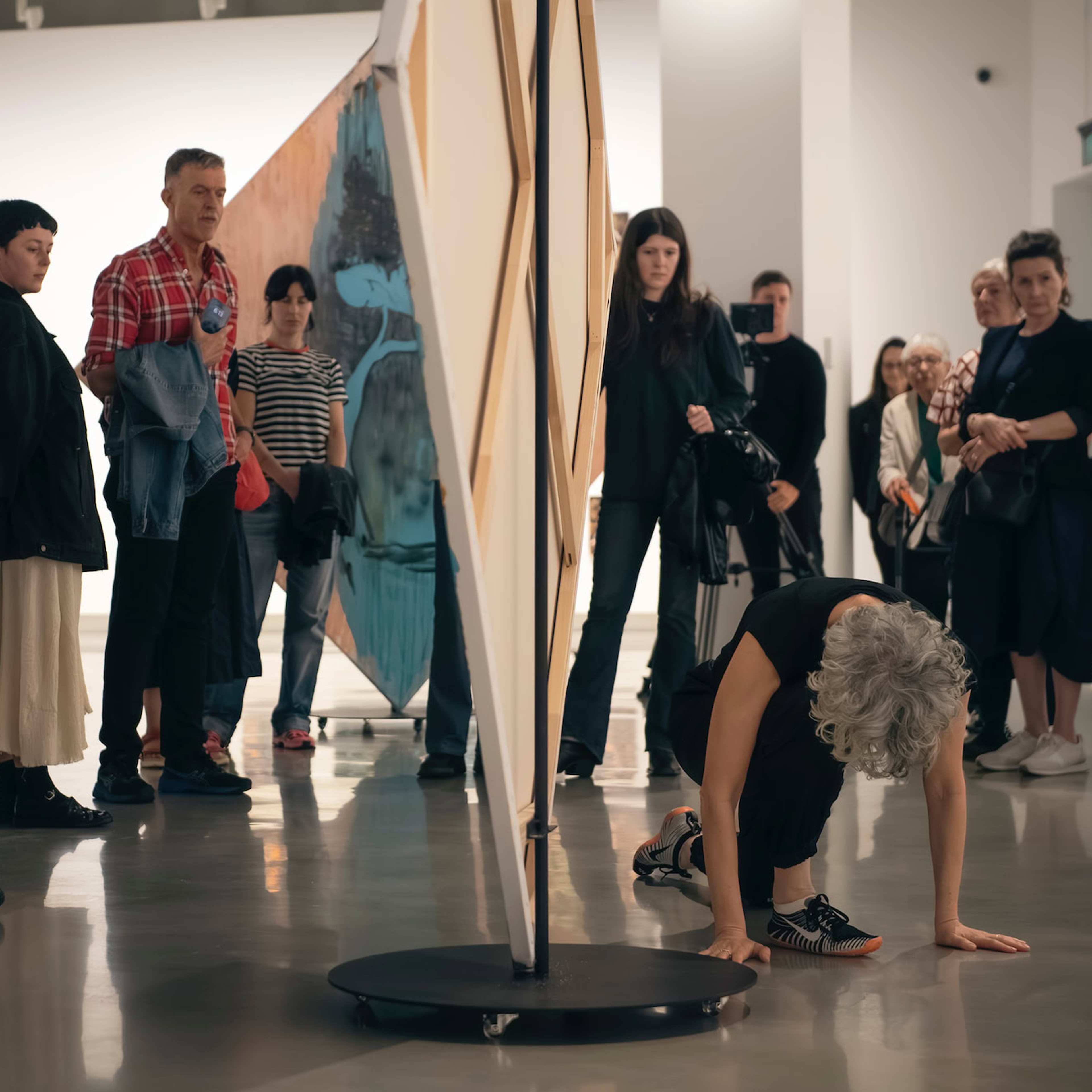 A dancer crouches on the floor during a performance art piece in a gallery, while a group of attentive people watch from around the room.