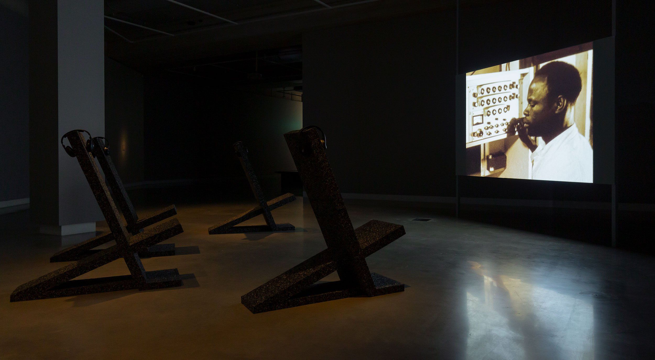 A dimly lit gallery features three angled black benches facing a large screen showing a man in front of a control panel. The floor is reflective, and headphones are attached to each bench.
