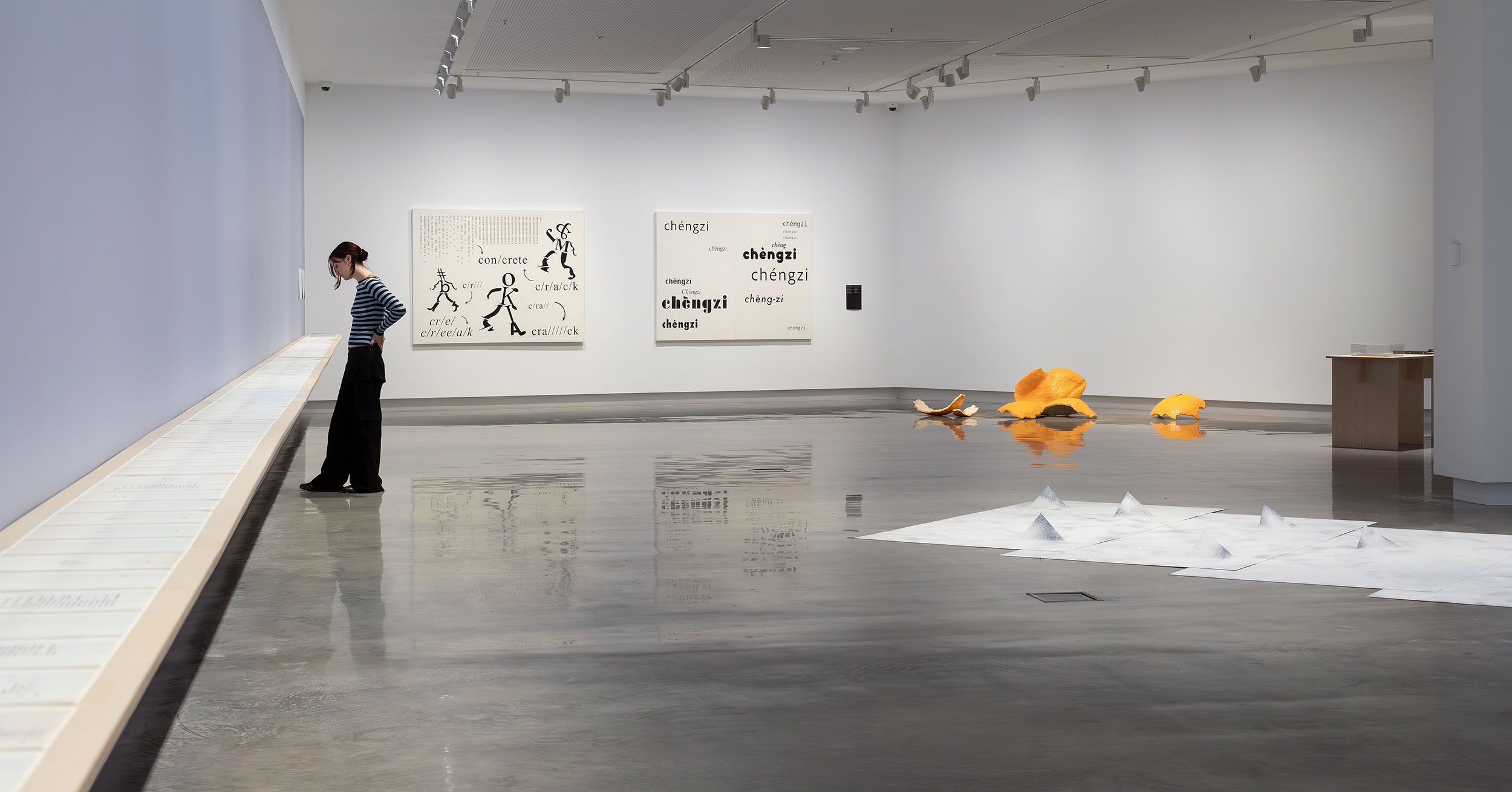 A person stands in a spacious, modern art gallery viewing a long, wall-mounted display. Artworks, including text-based pieces, sculptural objects, and paper installations, are arranged throughout the minimalist room.