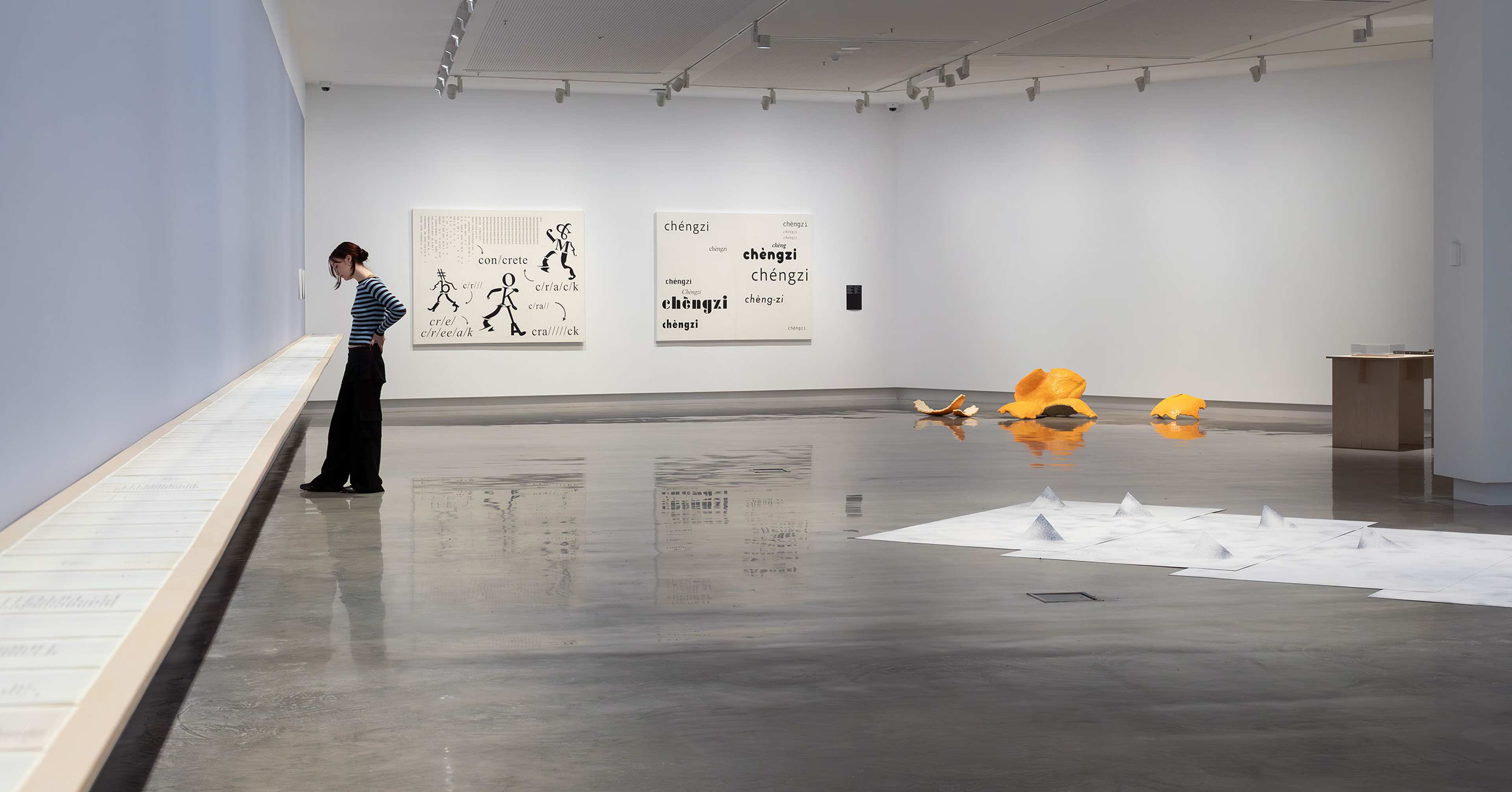 A person stands in a spacious, modern art gallery viewing a long, wall-mounted display. Artworks, including text-based pieces, sculptural objects, and paper installations, are arranged throughout the minimalist room.