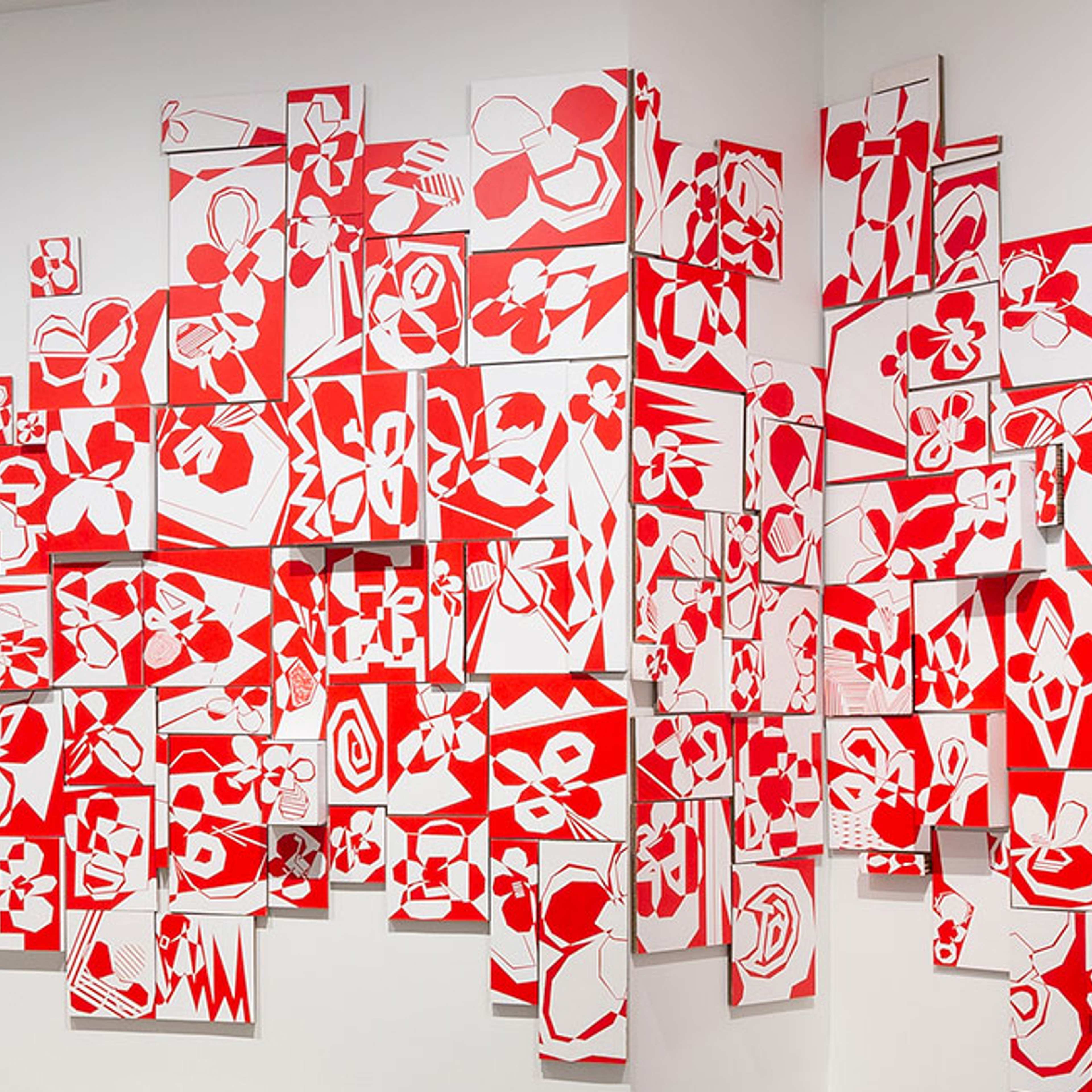 A wall-mounted art installation features numerous rectangular panels with bold red and white abstract floral and geometric designs, arranged in an overlapping, clustered pattern across a corner of a white gallery space.