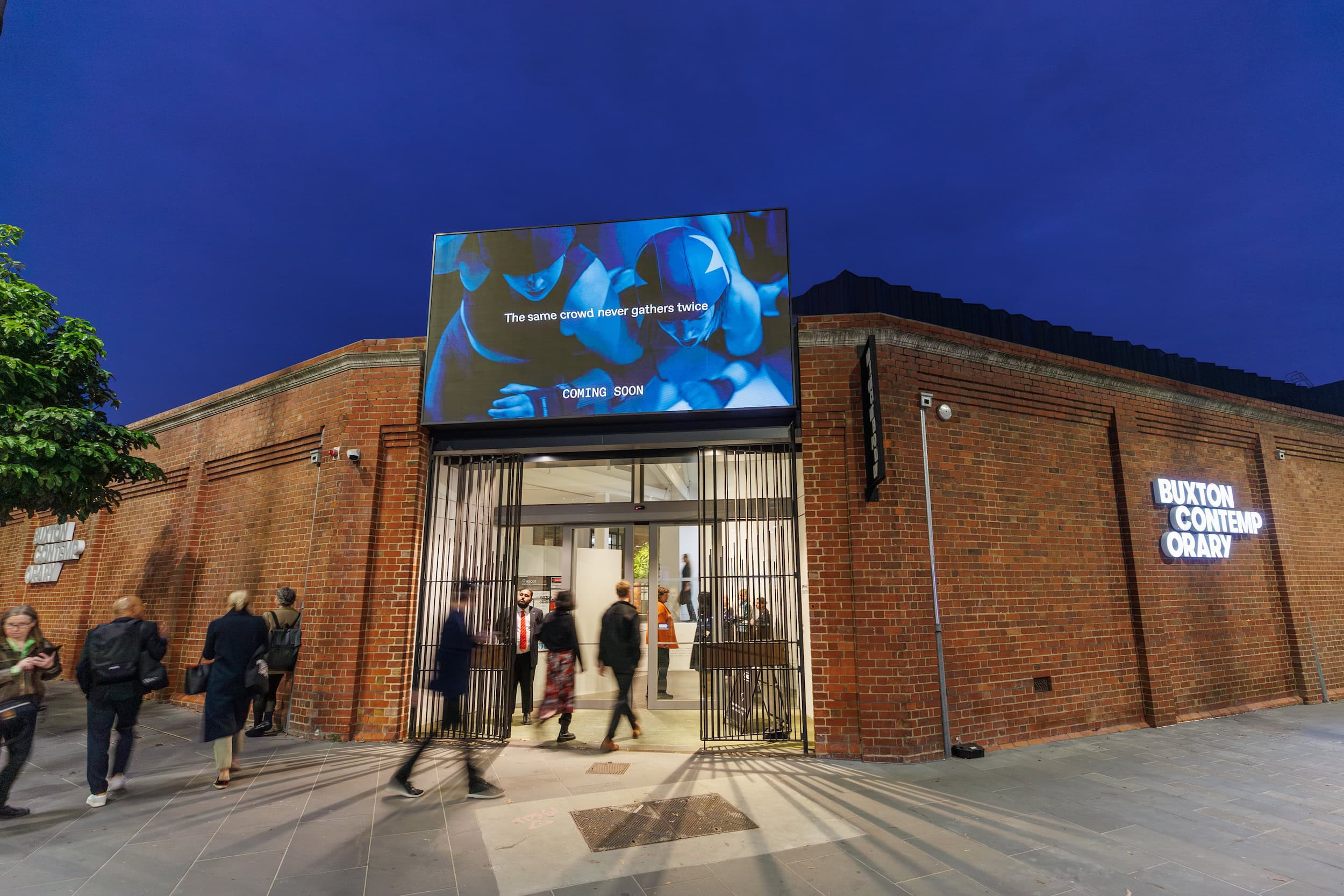 People enter and exit a modern brick art gallery at dusk. A digital billboard above the entrance reads, The same crowd never gathers twice. The building is labeled Buxton Contemporary with illuminated signage.