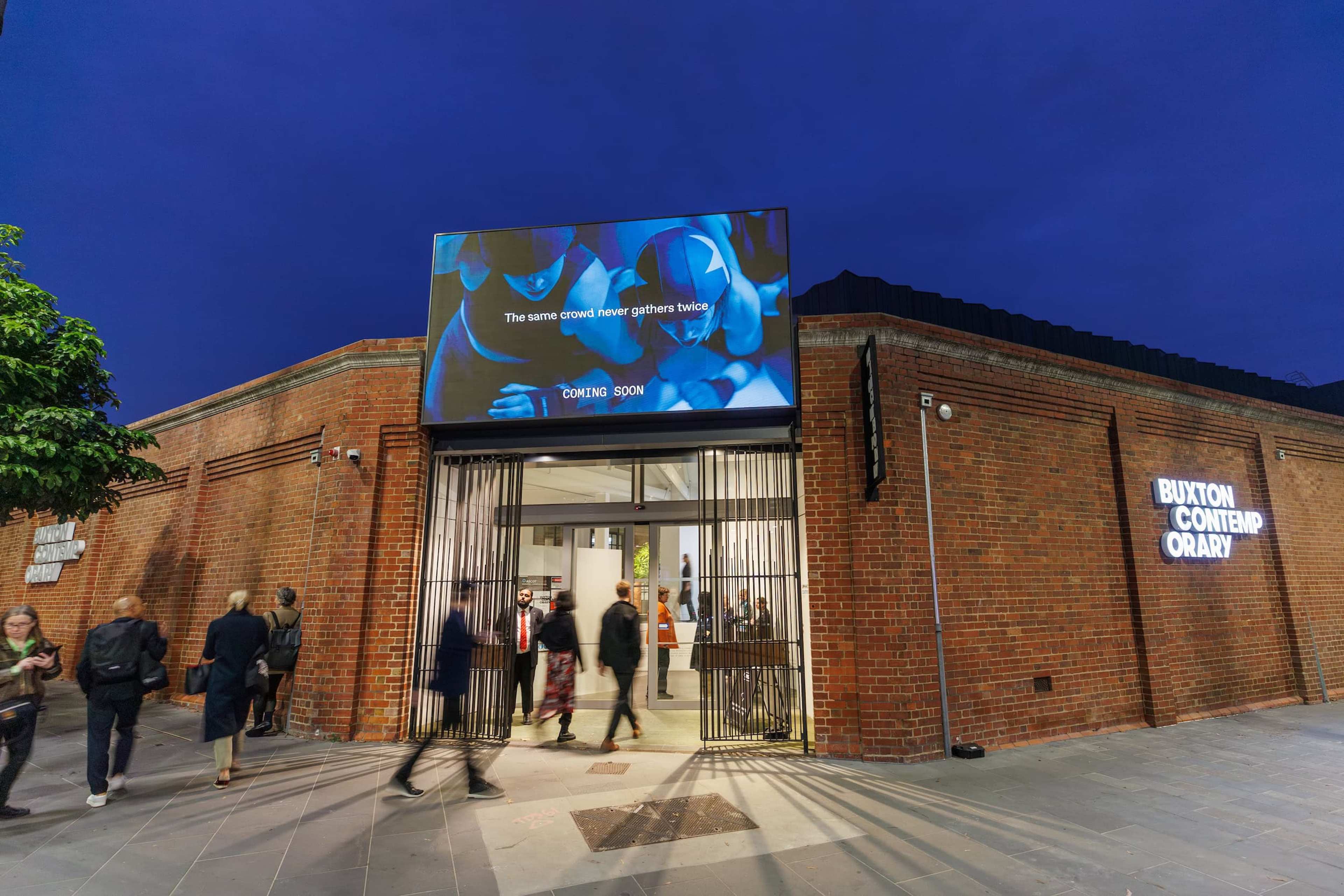 People enter and exit a modern brick art gallery at dusk. A digital billboard above the entrance reads, The same crowd never gathers twice. The building is labeled Buxton Contemporary with illuminated signage.