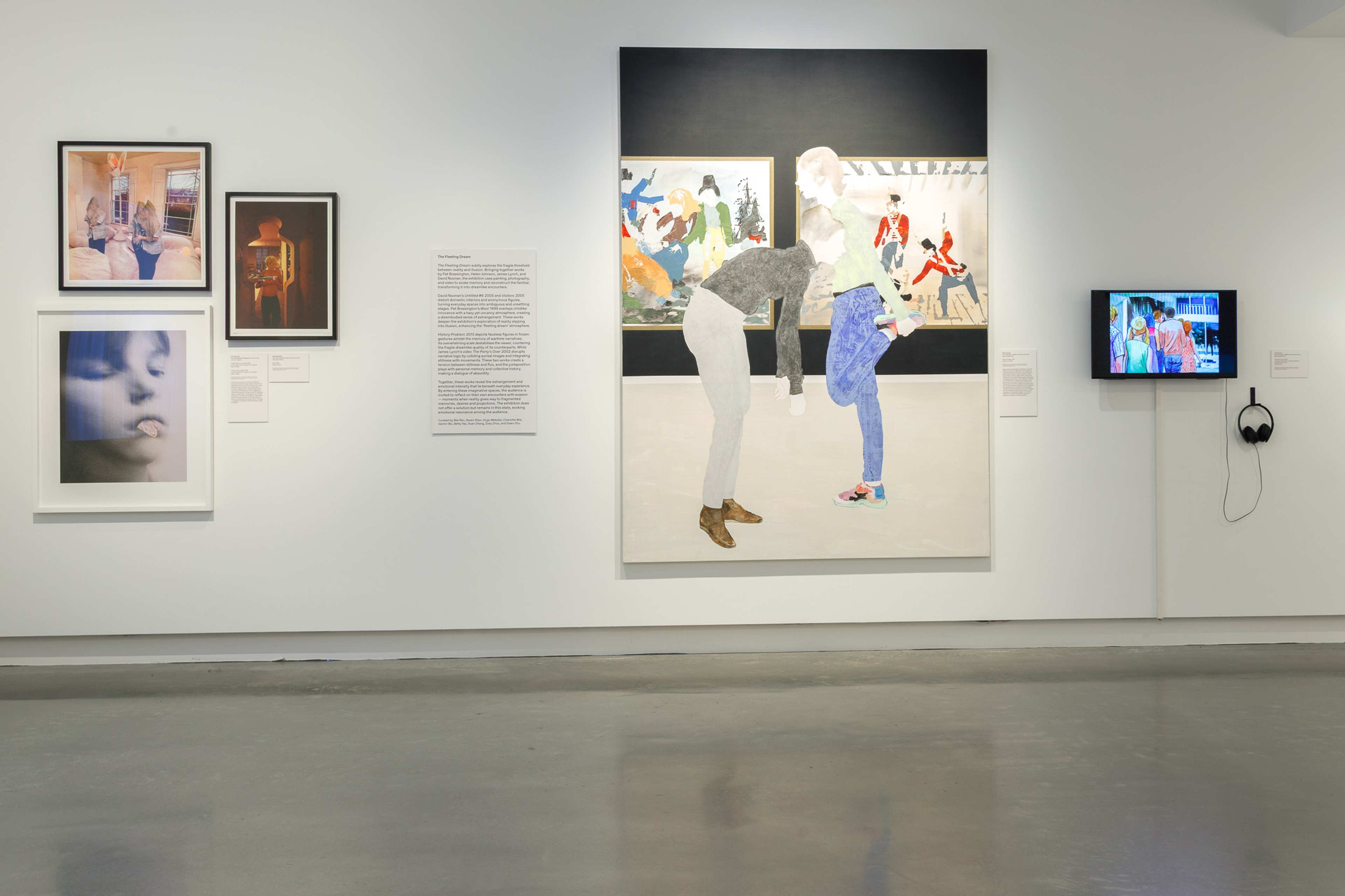 An art gallery wall displays framed photos, a large mixed-media painting of two figures in front of a mural, a wall text panel, and a monitor with headphones showing video art. The gallery floor is polished gray concrete.