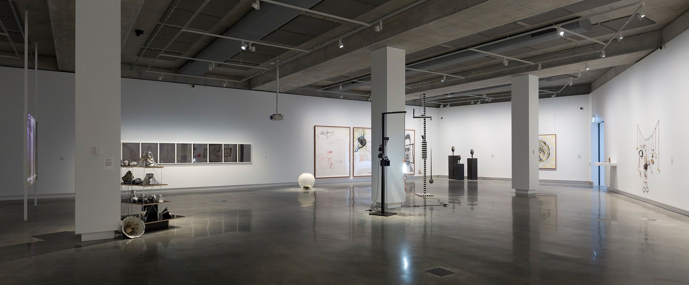 A spacious, modern art gallery with polished floors, white walls, and scattered sculptures and framed artworks displayed throughout the room under overhead lighting.