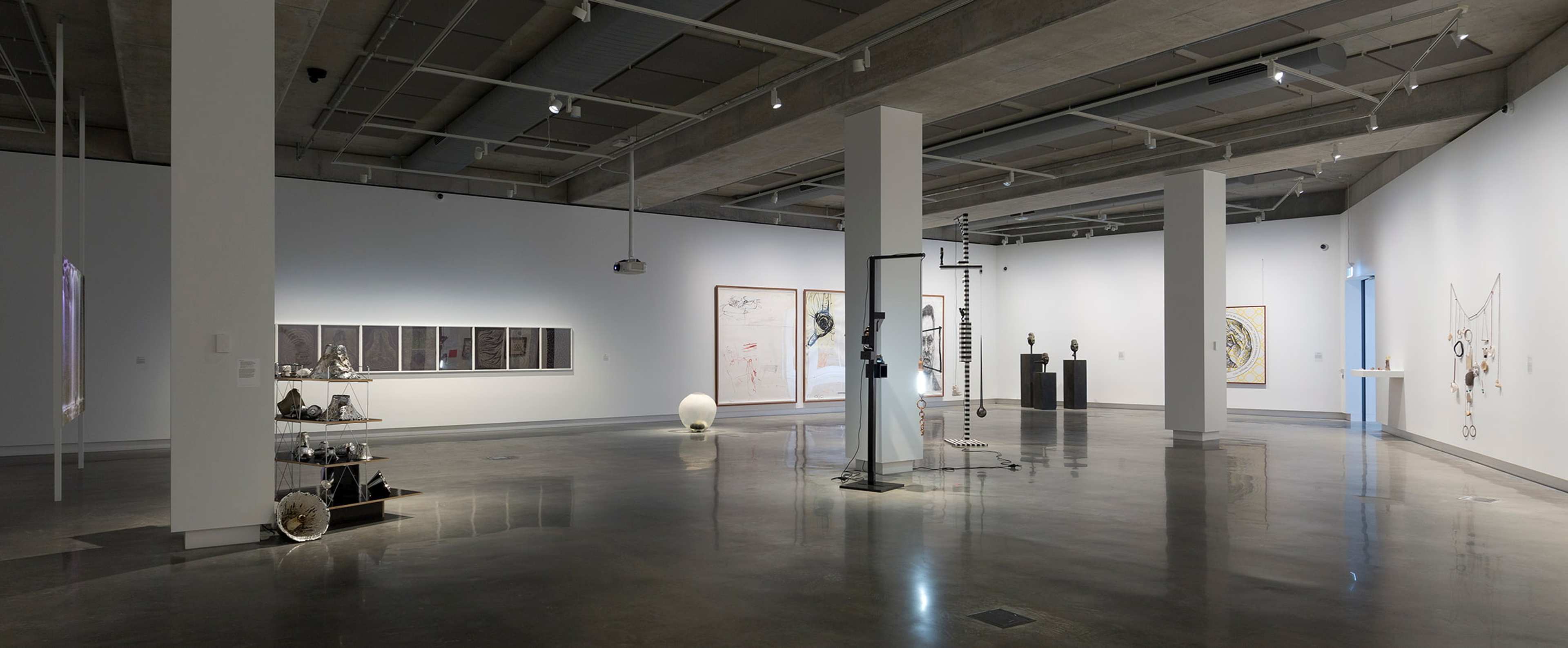 A spacious, modern art gallery with polished floors, white walls, and scattered sculptures and framed artworks displayed throughout the room under overhead lighting.