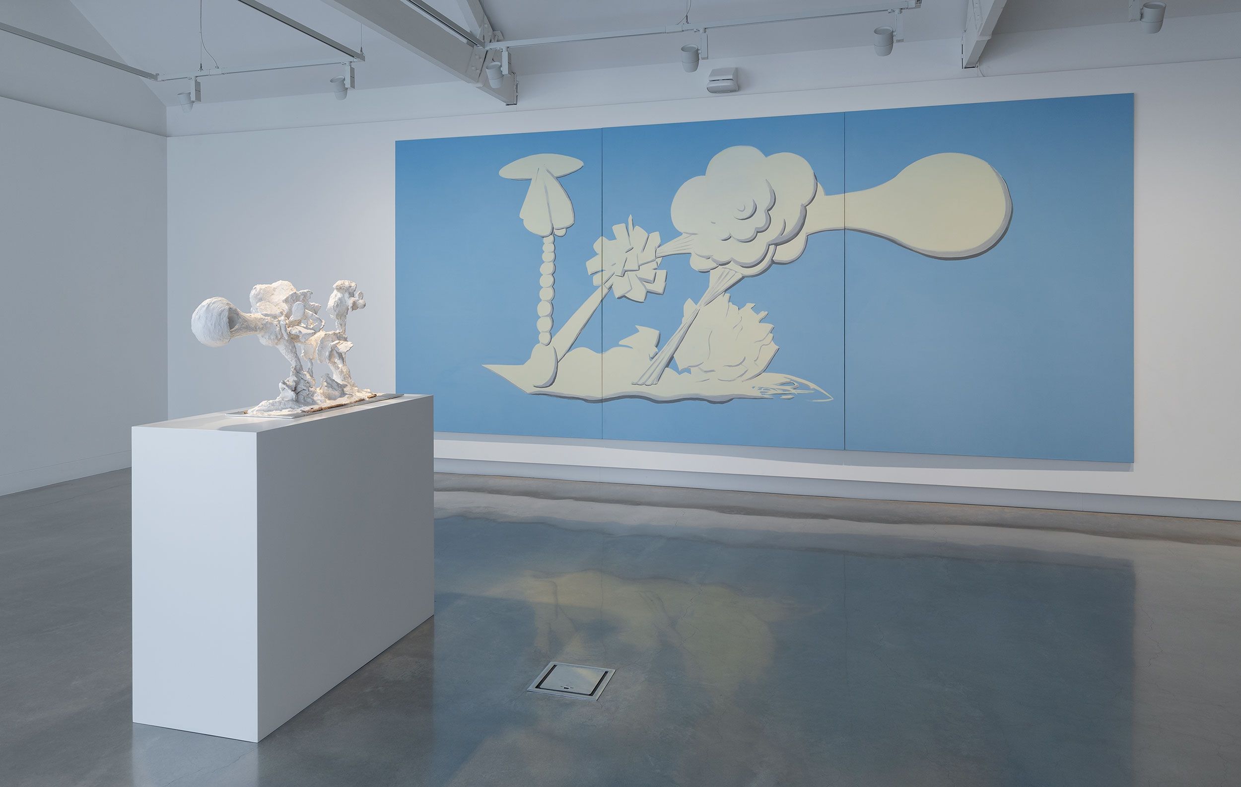 A white abstract sculpture on a pedestal stands in front of a large blue and cream artwork featuring cartoon-like cloud, smoke, and burst shapes, displayed in a modern, well-lit gallery space.