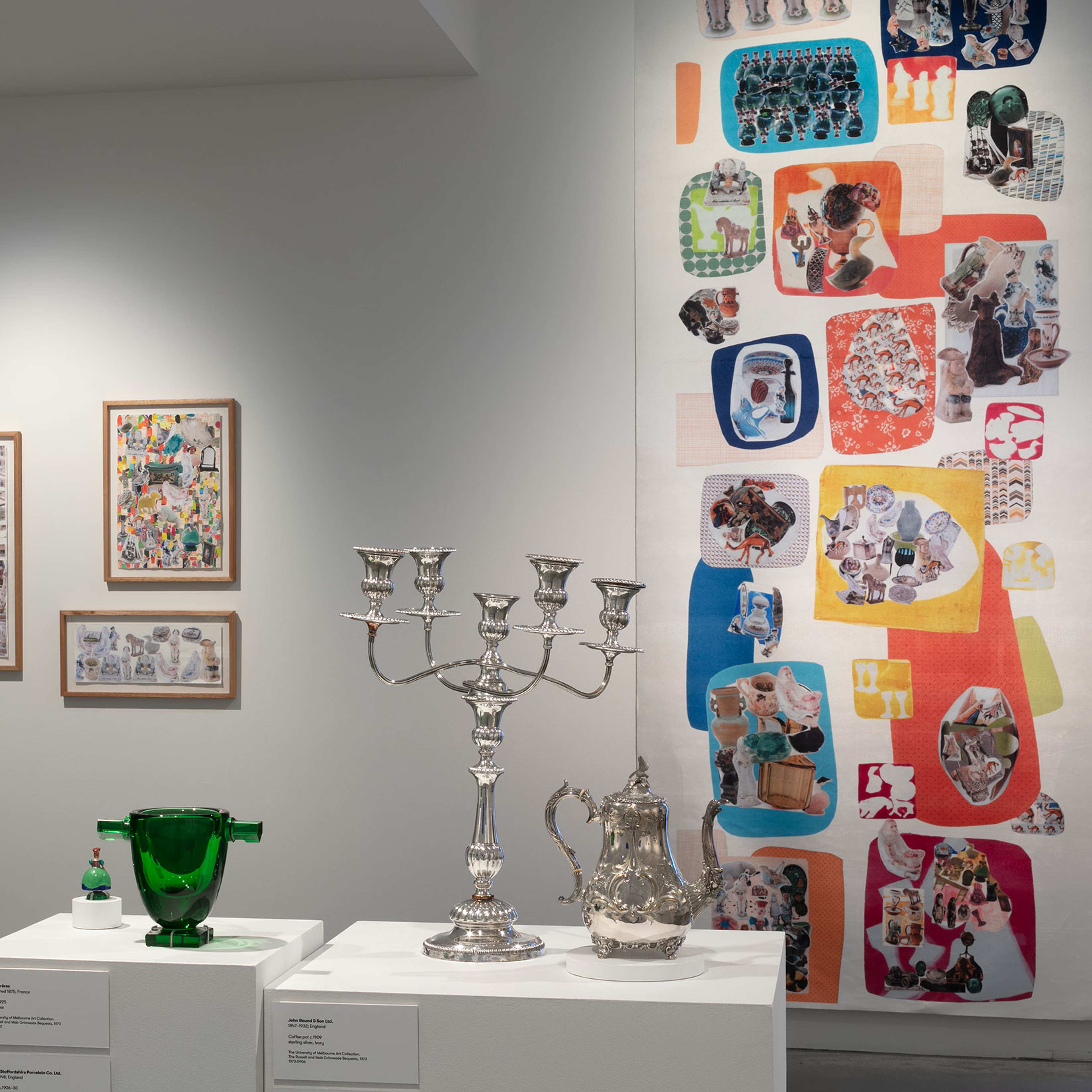 Gallery display with framed collage artworks on the wall, a large colorful textile piece, and glass and silver decorative objects including a candelabra and green vase on white pedestals.