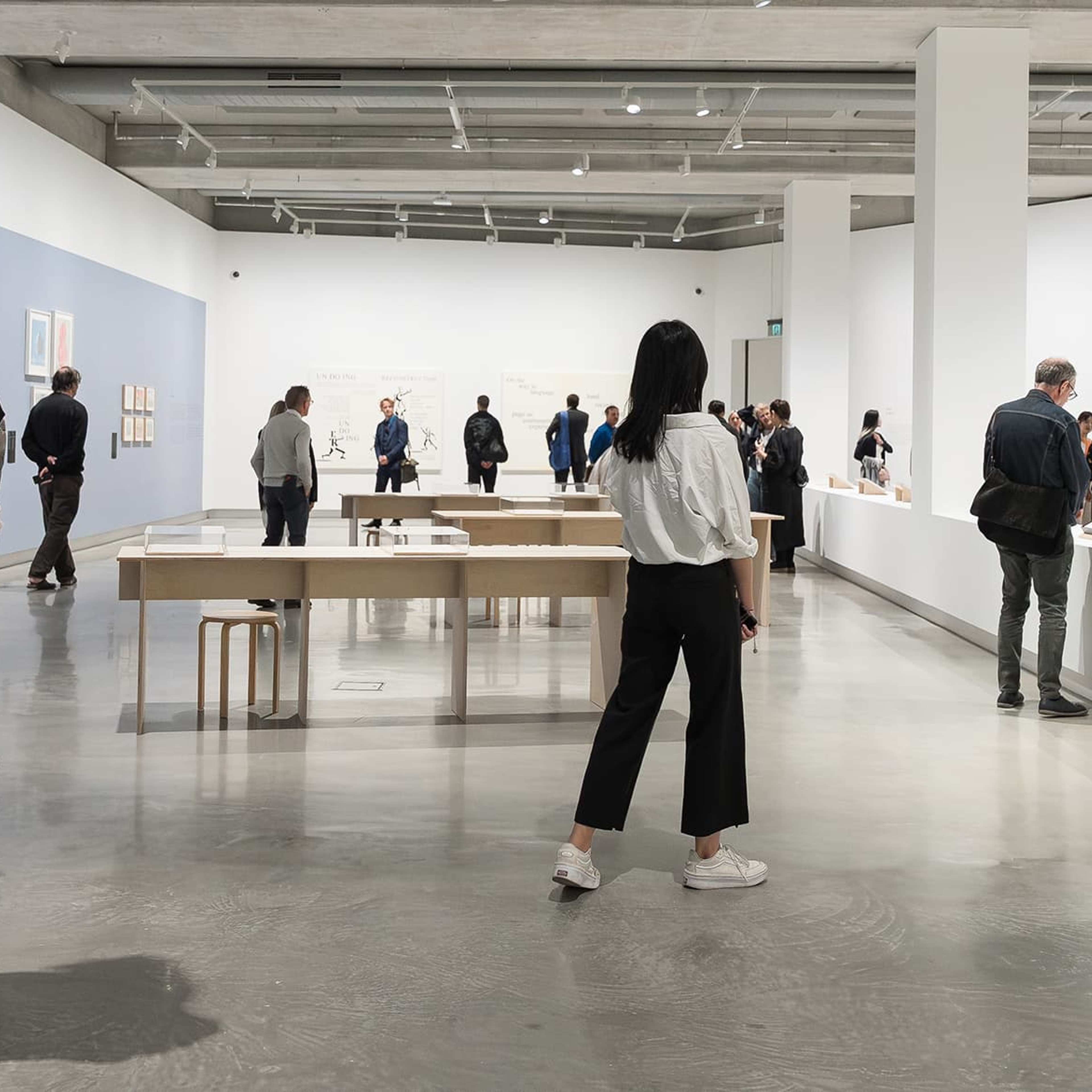 People walk around a spacious, modern art gallery with white walls, artwork, and display tables. Some view exhibits while others converse. The atmosphere is bright and quiet.