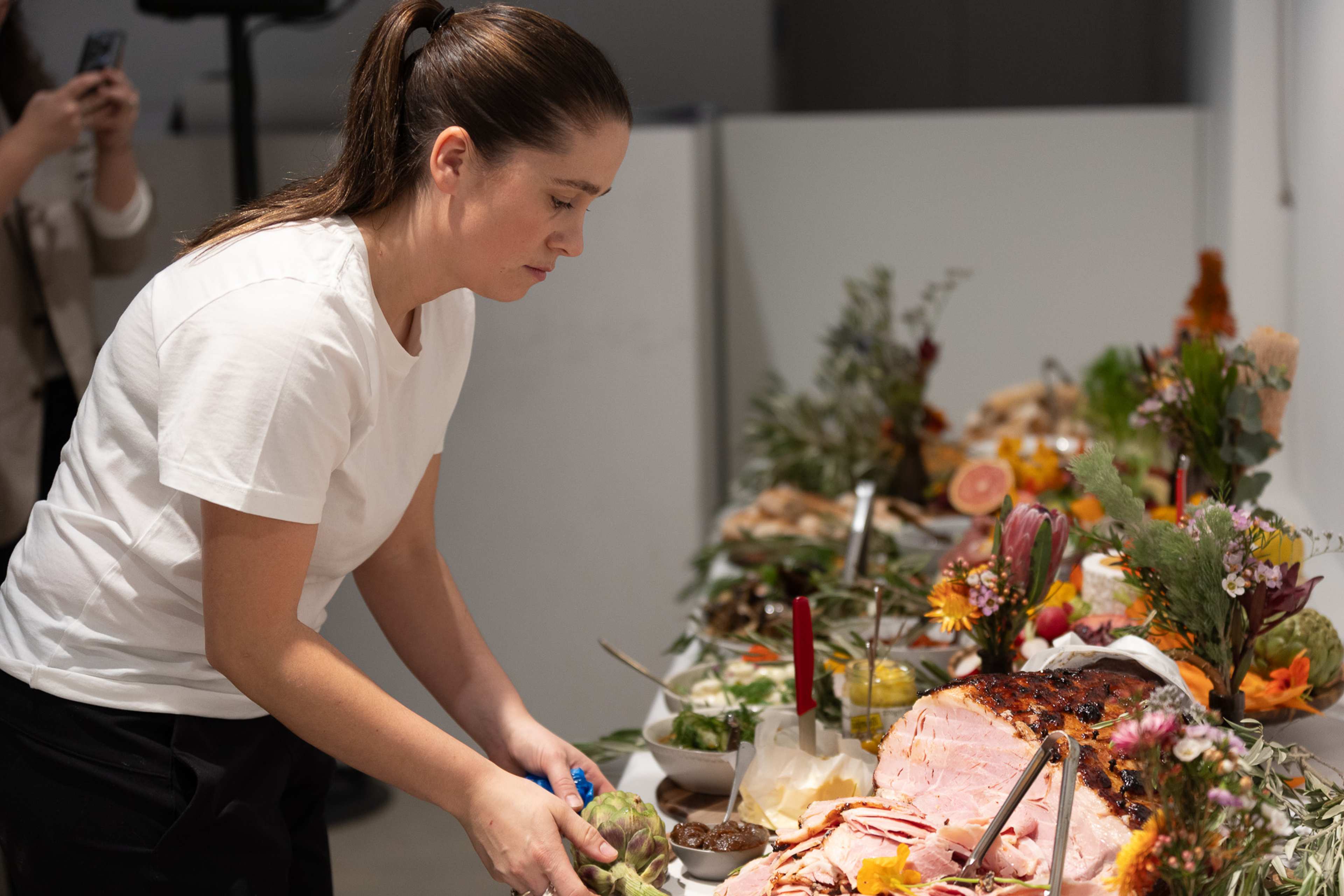 A person in a white t-shirt arranges food and decorative items on a table filled with various dishes, flowers, and candles, preparing for a event..