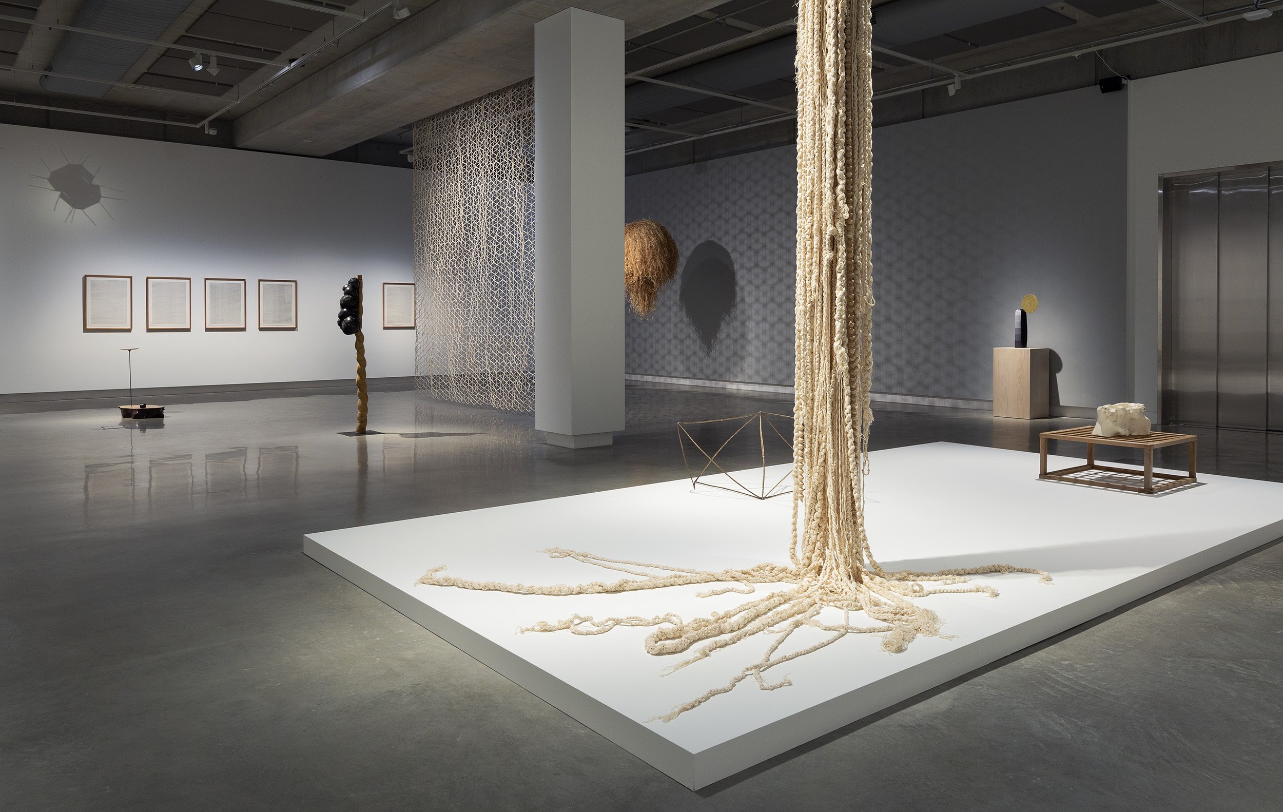 A modern art gallery with sculptures and installations, including hanging ropes on a white platform, wall-mounted artworks, abstract freestanding pieces, and geometric objects in a spacious, well-lit room with polished concrete floors.