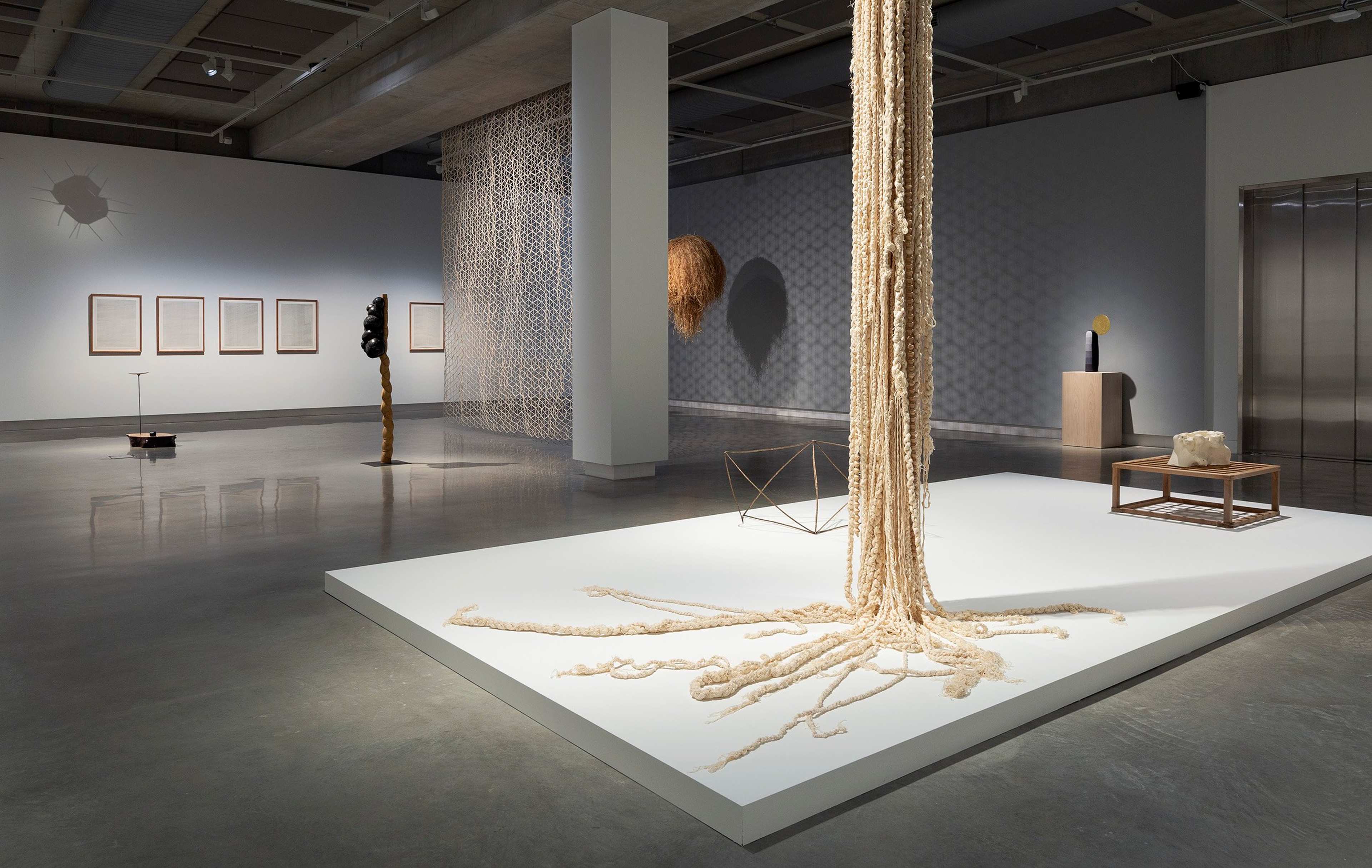 A modern art gallery with sculptures and installations, including hanging ropes on a white platform, wall-mounted artworks, abstract freestanding pieces, and geometric objects in a spacious, well-lit room with polished concrete floors.