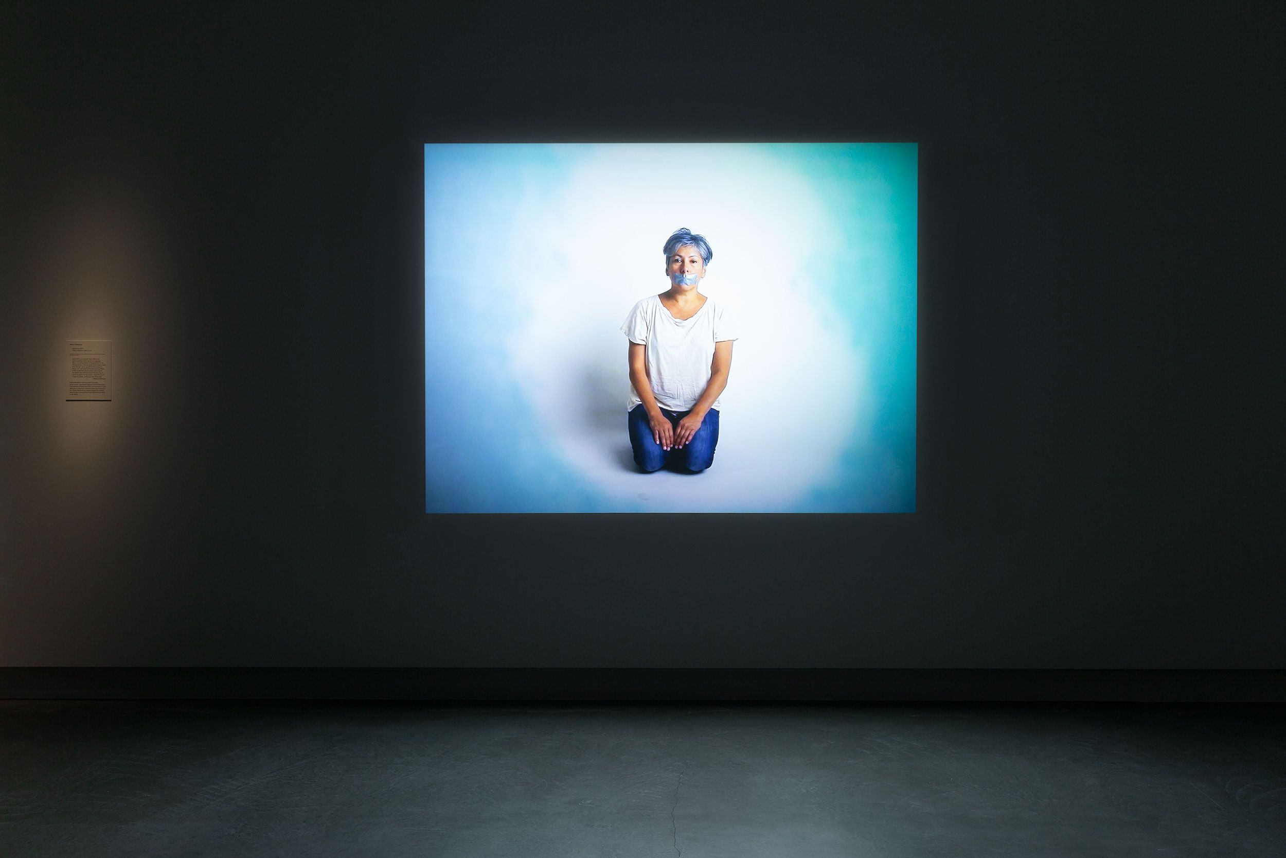 A screen with a person in white t-shirt kneeling in a white room with tape over their mouth in a dark gallery