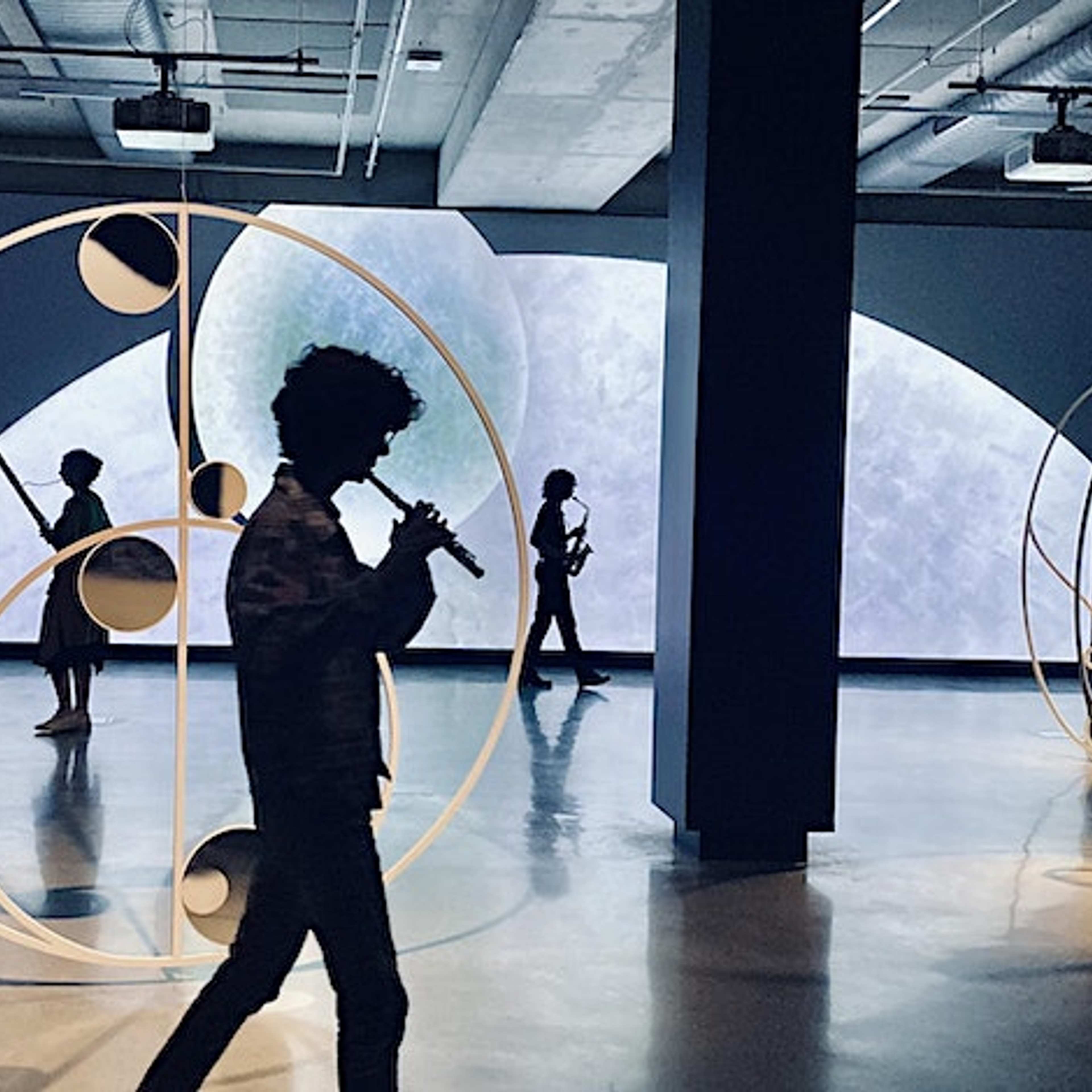 Silhouetted people walk among large circular sculptures in a dimly lit, modern indoor space with a large abstract display screen glowing in the background.