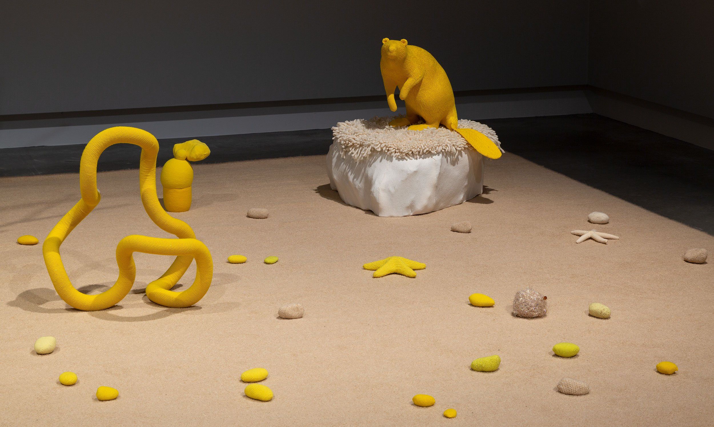 A yellow sculpture of an animal, possibly a beaver, sits on a white platform surrounded by abstract yellow shapes and stones scattered on a sandy floor in a minimalist gallery setting.