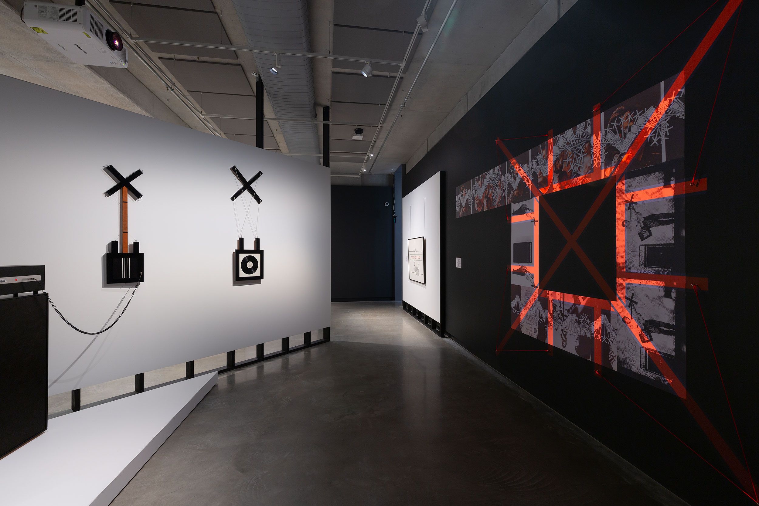 A modern art gallery features abstract wall-mounted sculptures on the left and a large geometric mixed-media piece with red tape and monochrome prints on the right, set in a sleek space with concrete floors and spot lighting.