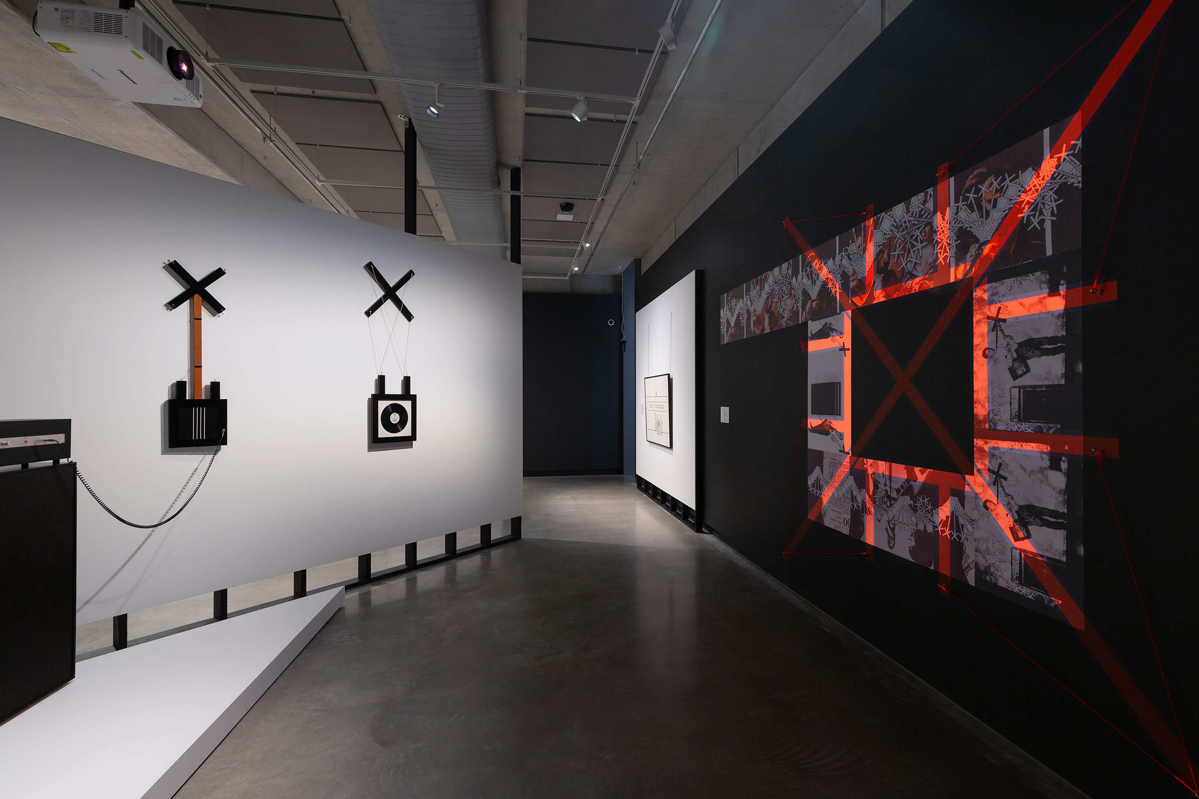A modern art gallery features abstract wall-mounted sculptures on the left and a large geometric mixed-media piece with red tape and monochrome prints on the right, set in a sleek space with concrete floors and spot lighting.