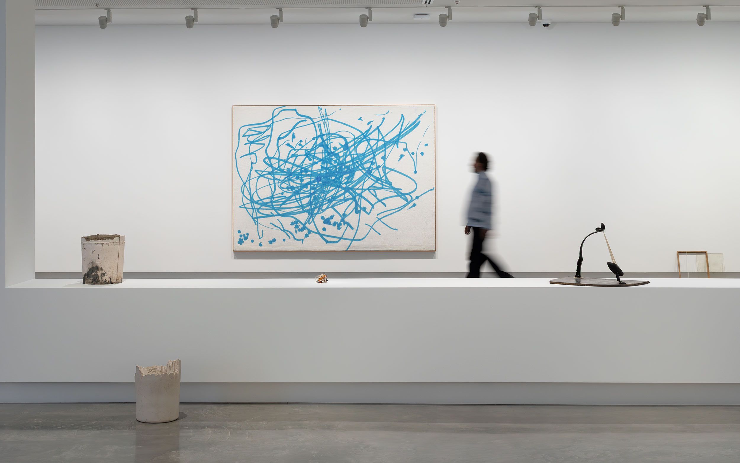 A person examines a blue and white scribble painting in a modern, spacious art gallery with polished floors. 