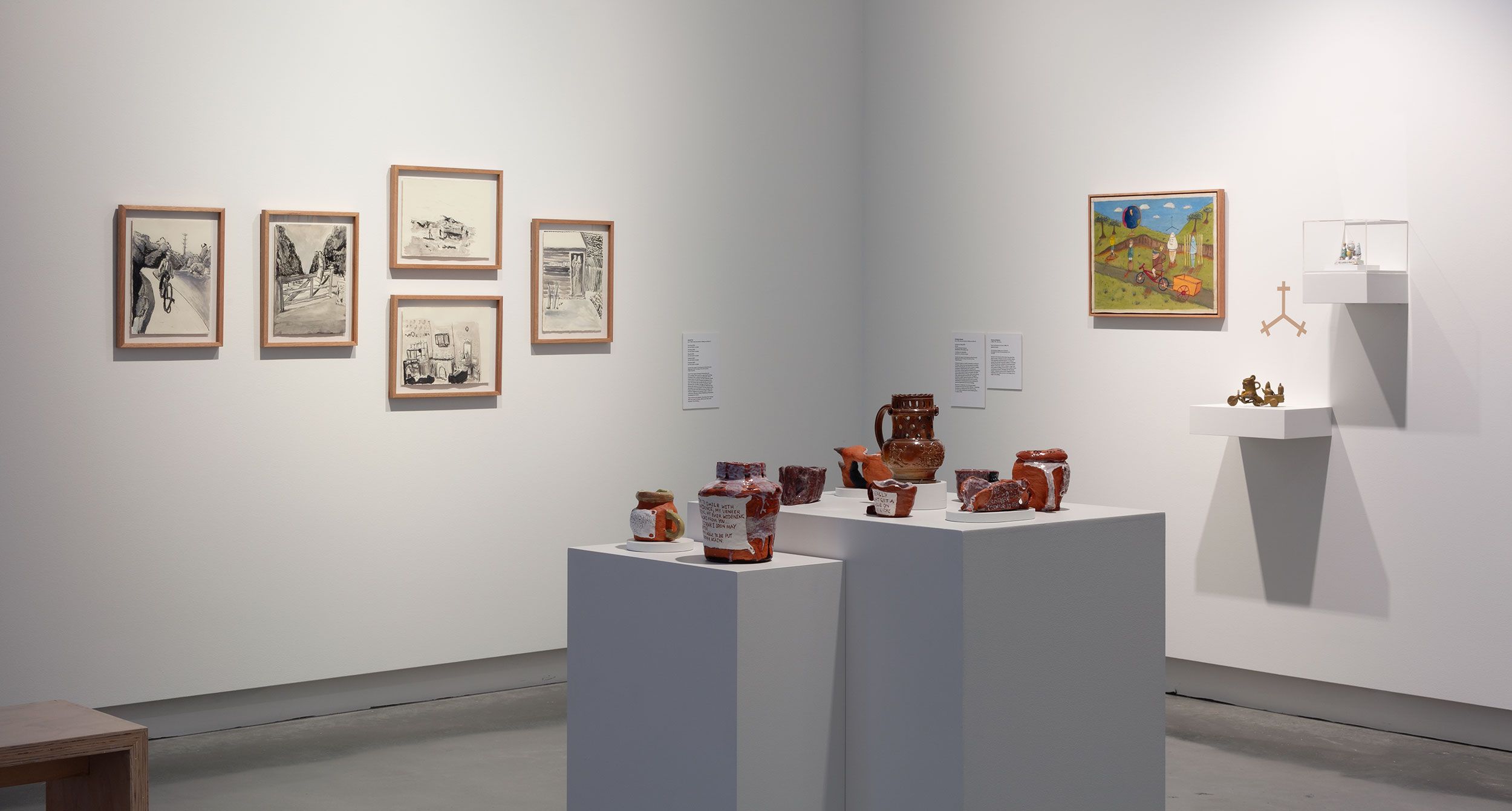A gallery exhibit with framed drawings and a colorful landscape painting on white walls, plus ceramic vessels and sculptures displayed on white pedestals and shelves. The space is brightly lit and minimalist.