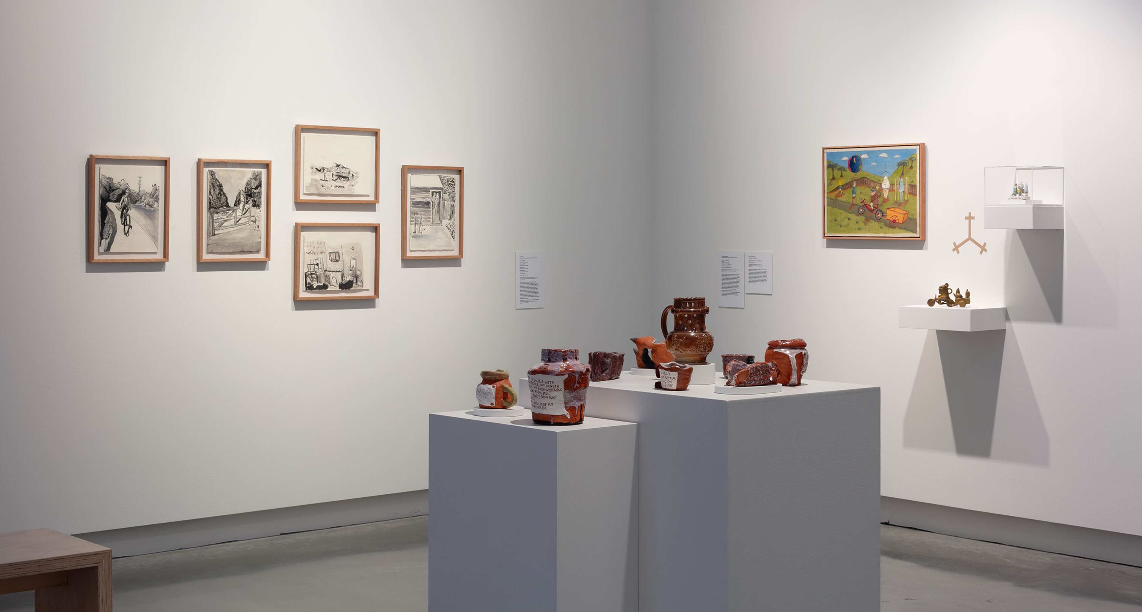 A gallery exhibit with framed drawings and a colorful landscape painting on white walls, plus ceramic vessels and sculptures displayed on white pedestals and shelves. The space is brightly lit and minimalist.