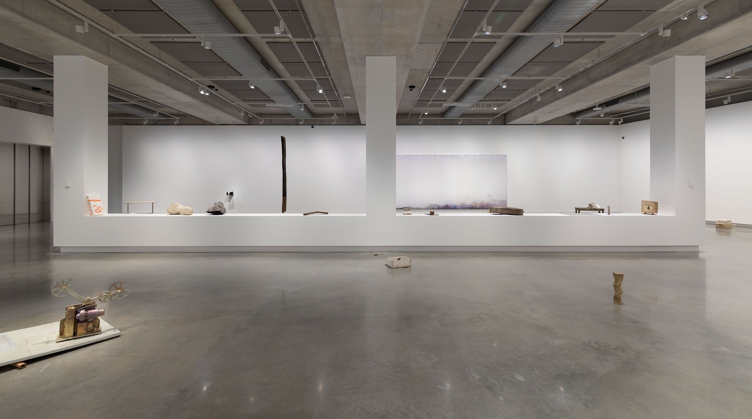 A contemporary art gallery with minimal sculptures: two organic-shaped stones on a white pedestal, a tall vertical sculpture in the background, a wall-mounted piece, and an abstract artwork on the far right wall.