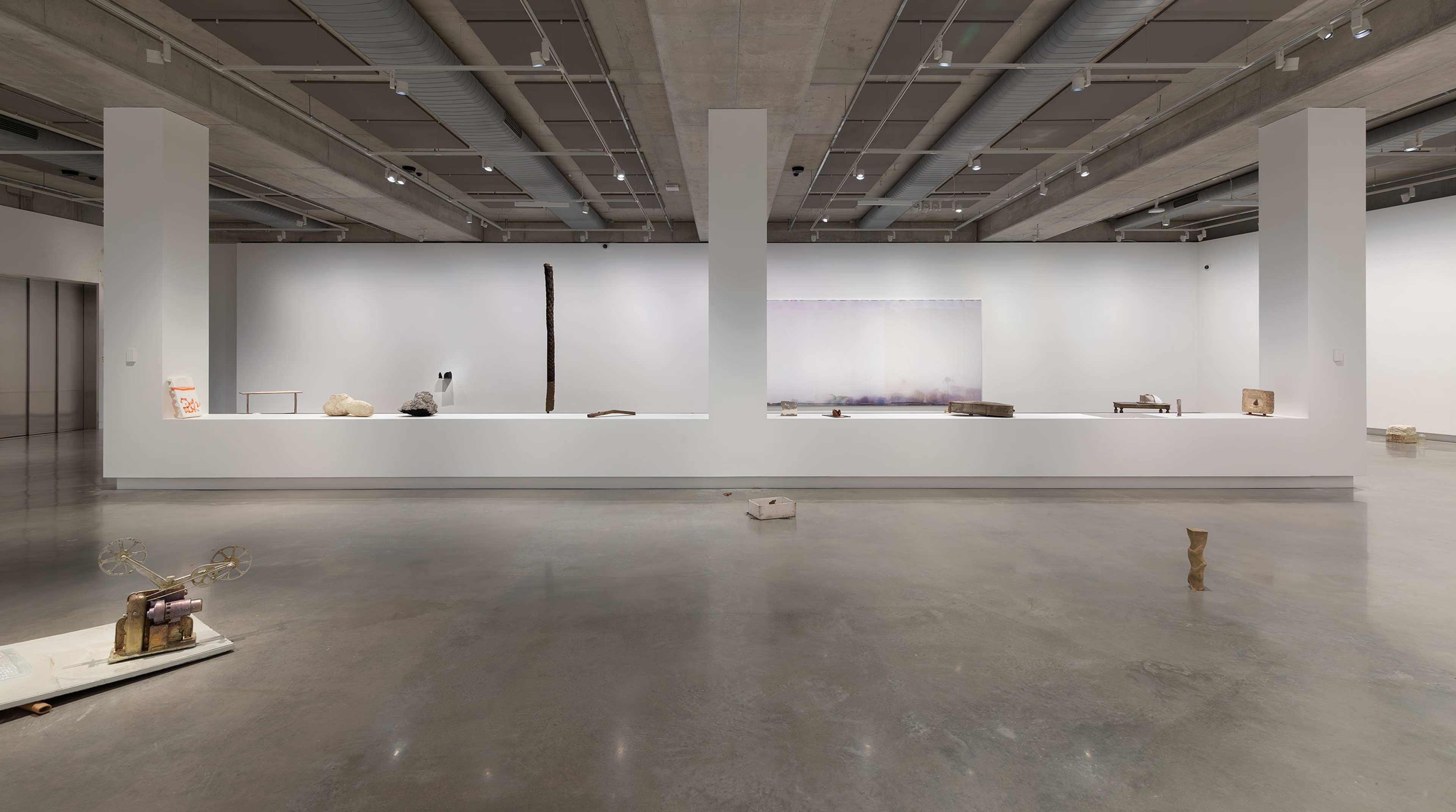 A contemporary art gallery with minimal sculptures: two organic-shaped stones on a white pedestal, a tall vertical sculpture in the background, a wall-mounted piece, and an abstract artwork on the far right wall.