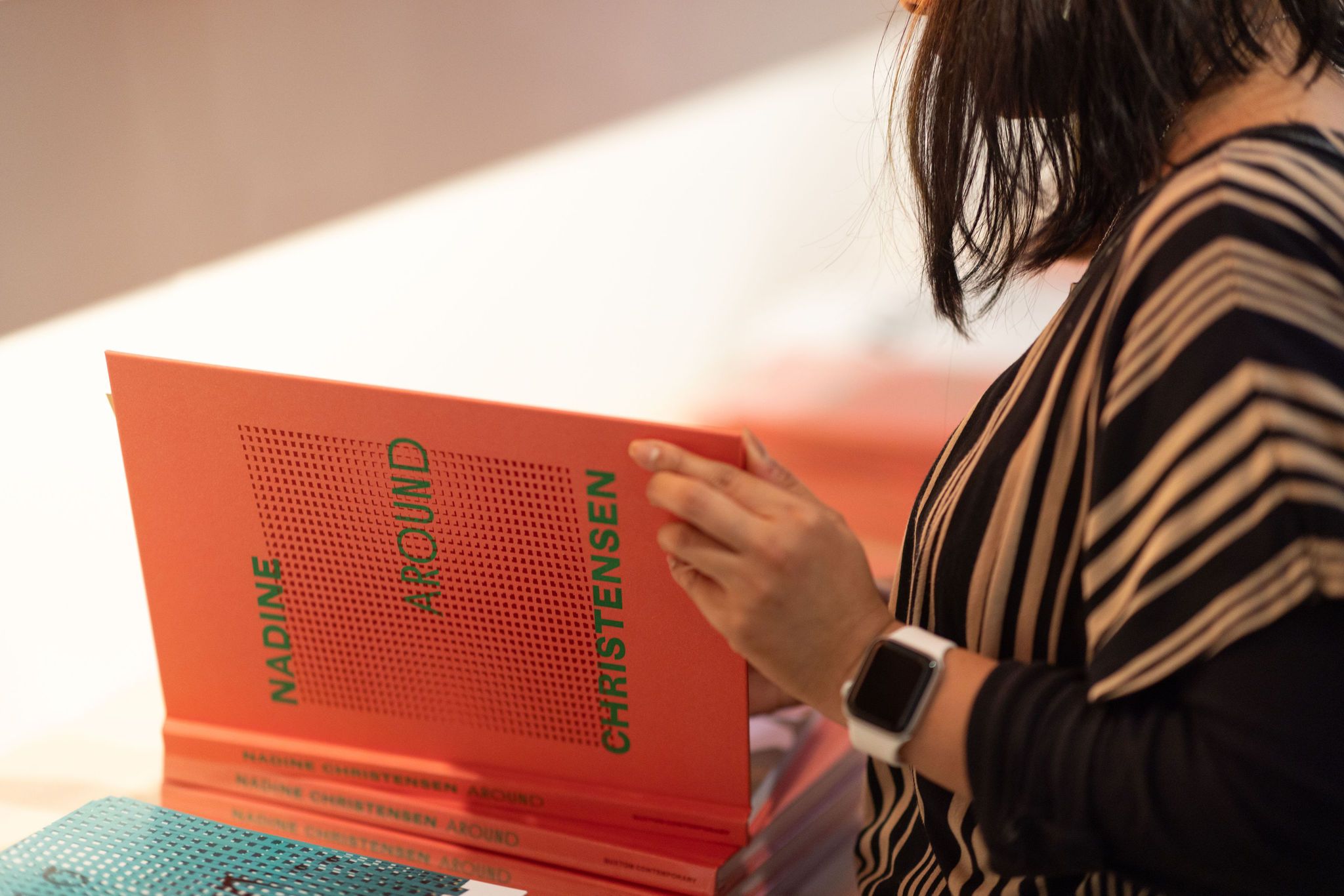 A person wearing a striped shirt and a smartwatch flips through a bright orange book titled Nadine Christensen Around, with more copies stacked on the table.