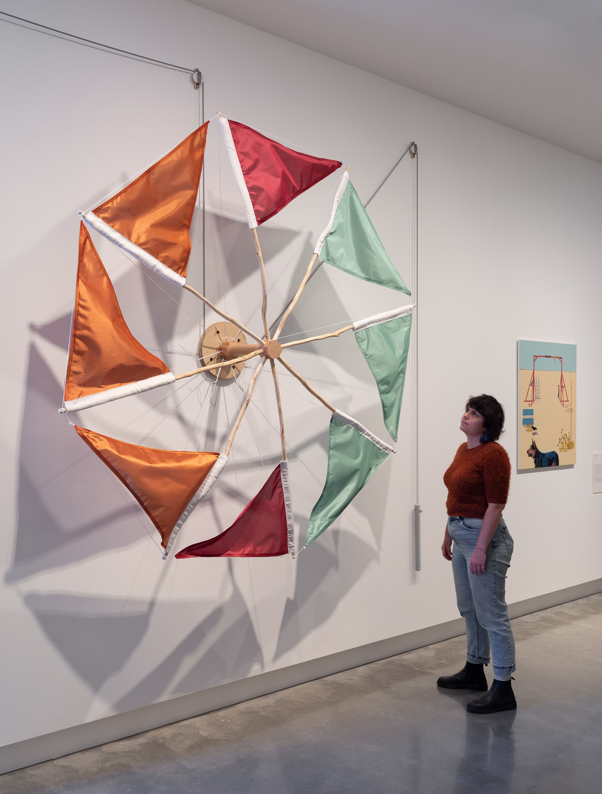 A person stands in front of a large, colorful geometric art installation made of red, orange, and light green fabric panels stretched on a wooden frame, mounted on a white gallery wall.