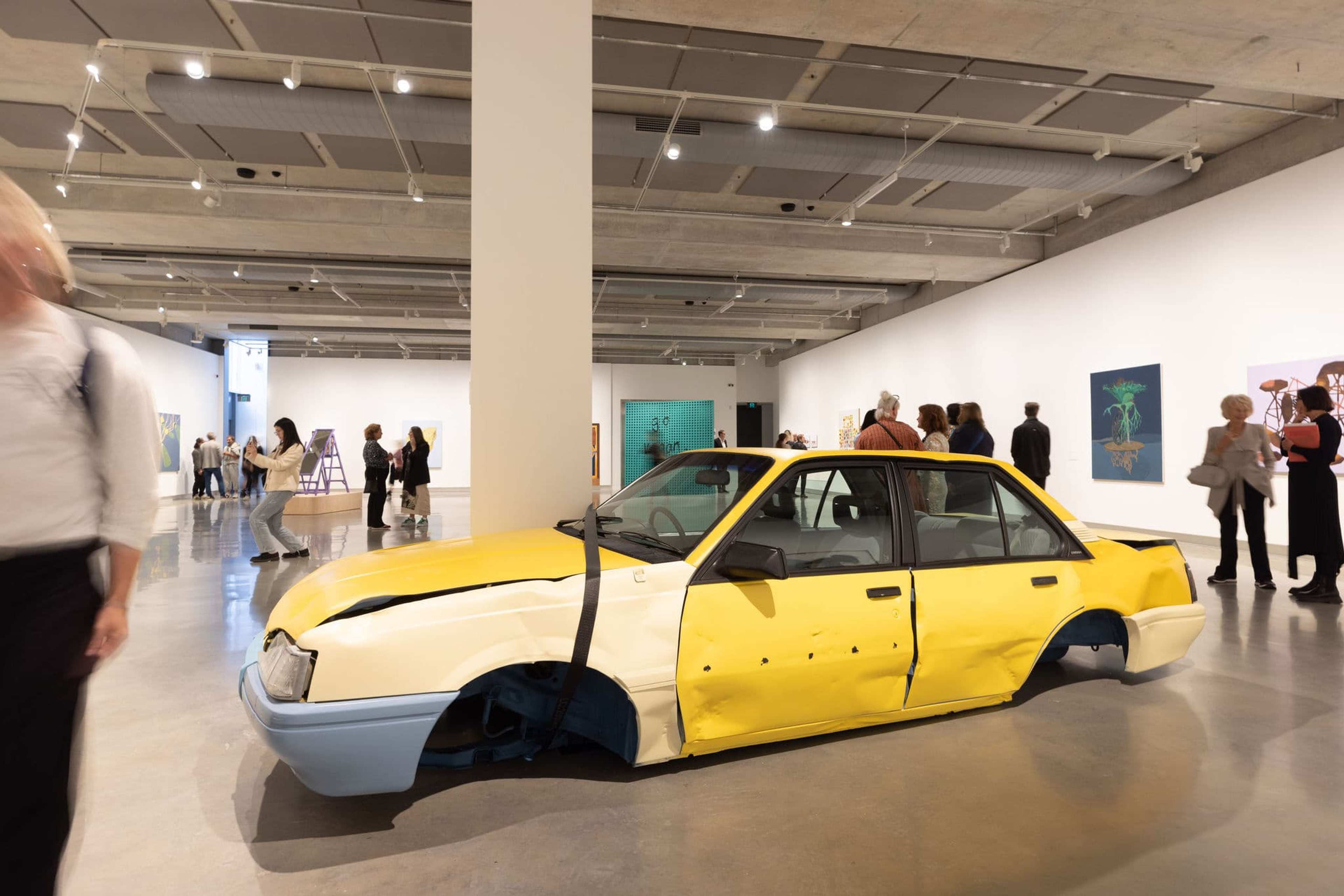 A yellow car with blue bumpers, squashed flat and missing wheels, is displayed as art in a bright modern gallery. People walk around and observe various artworks on the walls.
