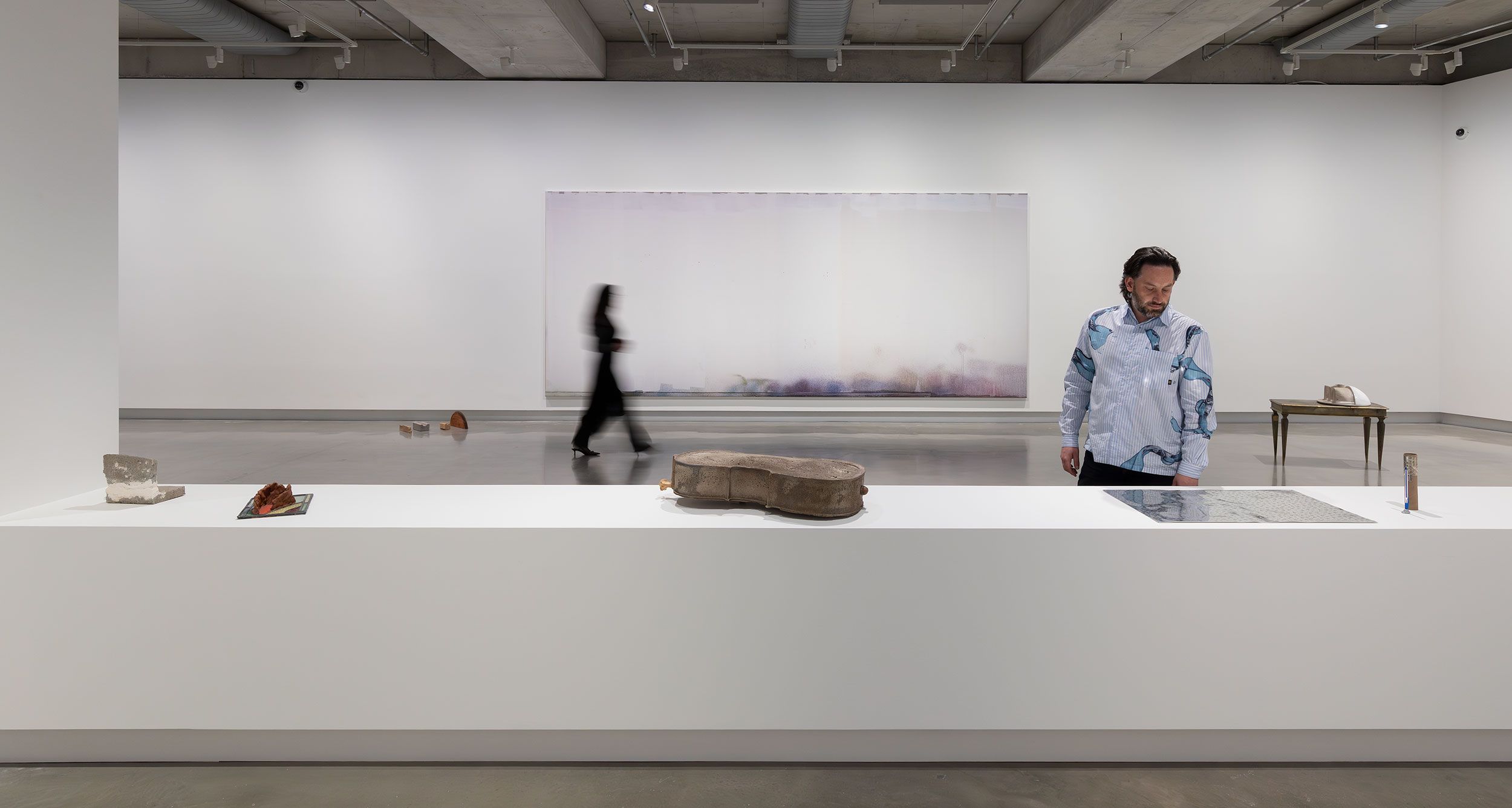 A person stands near a white display counter with sculptures and objects, while a blurred figure walks by in a modern, minimalist art gallery with a large abstract painting on the far wall.