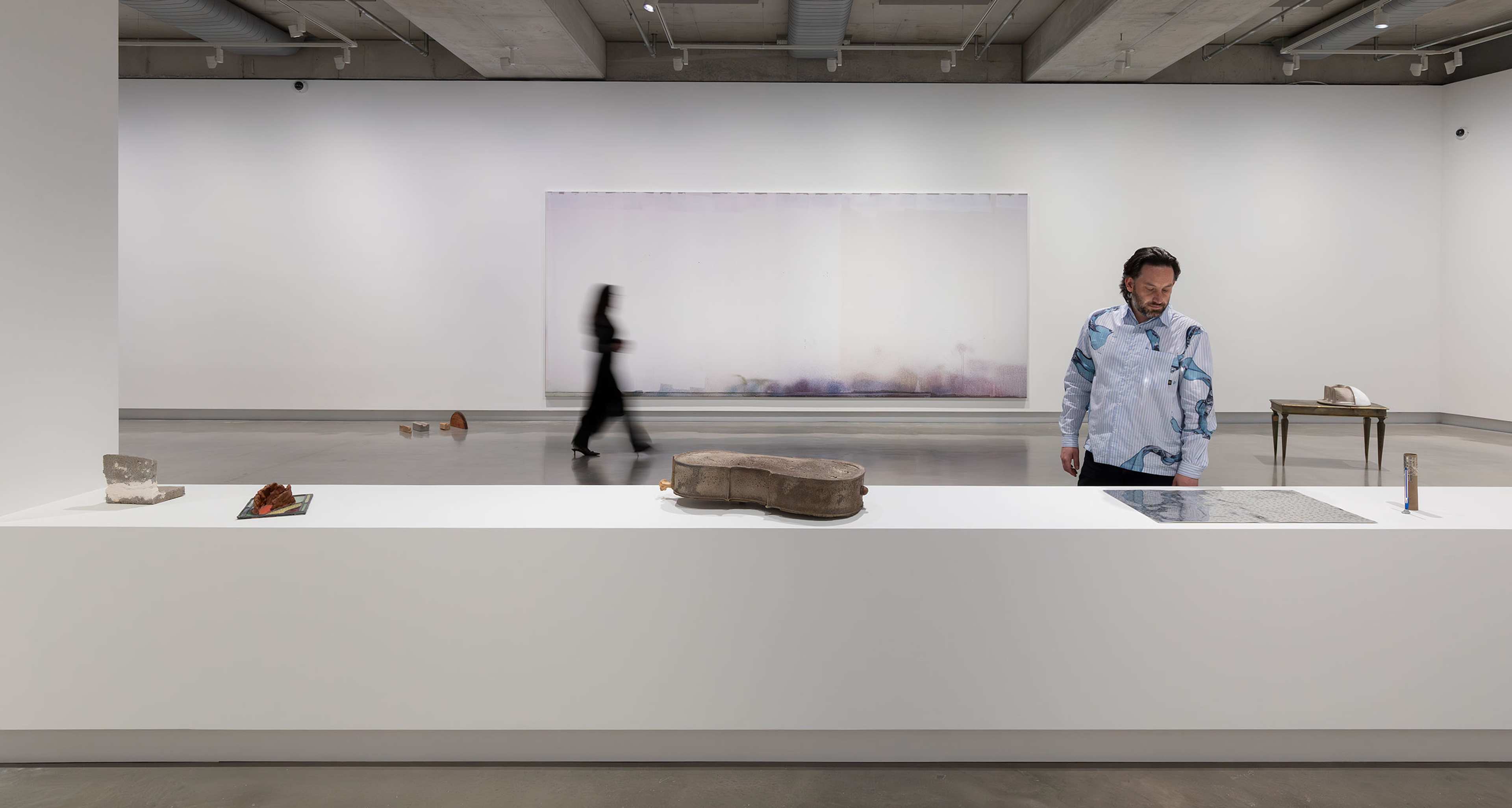 A person stands near a white display counter with sculptures and objects, while a blurred figure walks by in a modern, minimalist art gallery with a large abstract painting on the far wall.