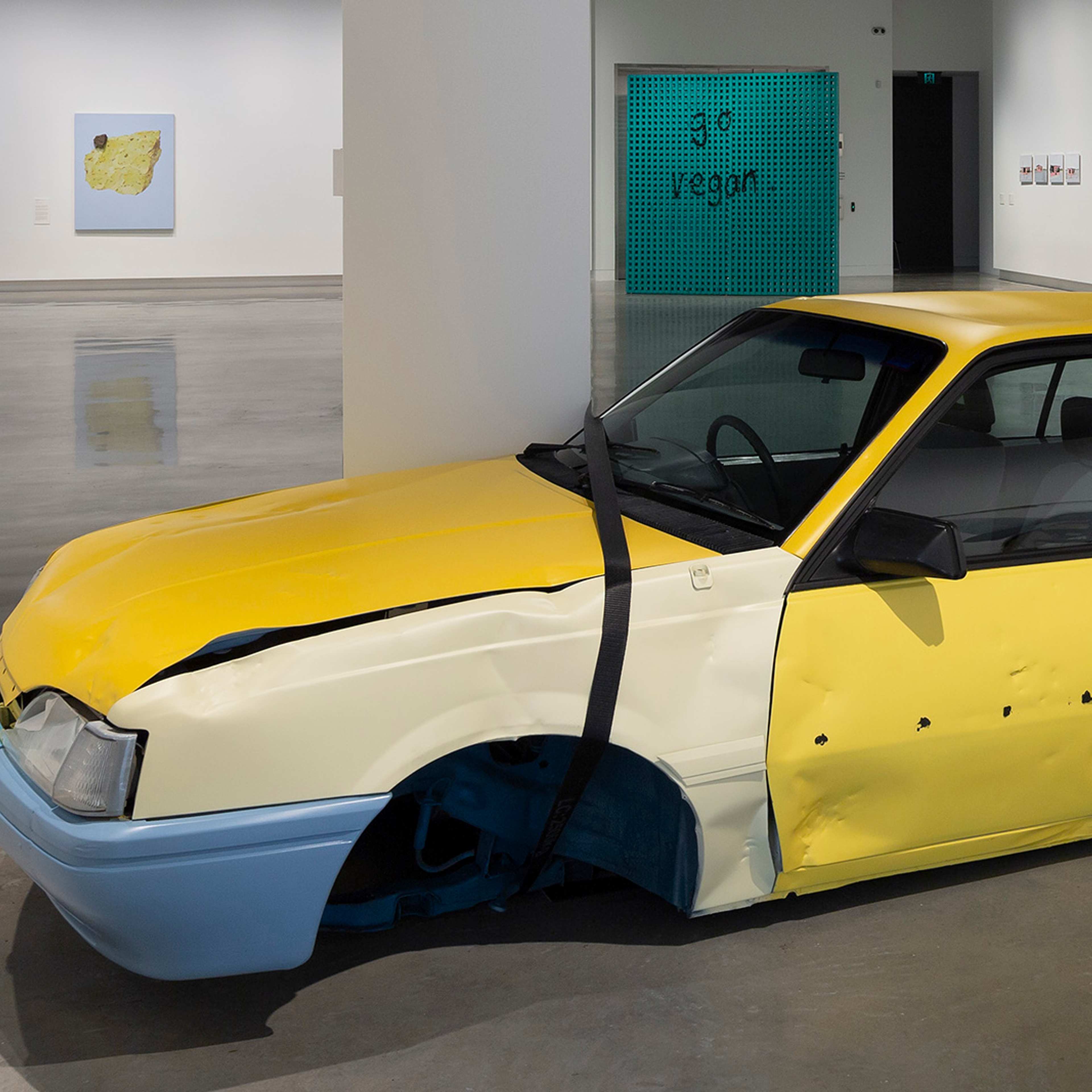 A yellow car with blue bumpers, heavily damaged and flattened, is displayed as an art installation in a modern gallery. The gallery has white walls and a polished concrete floor.