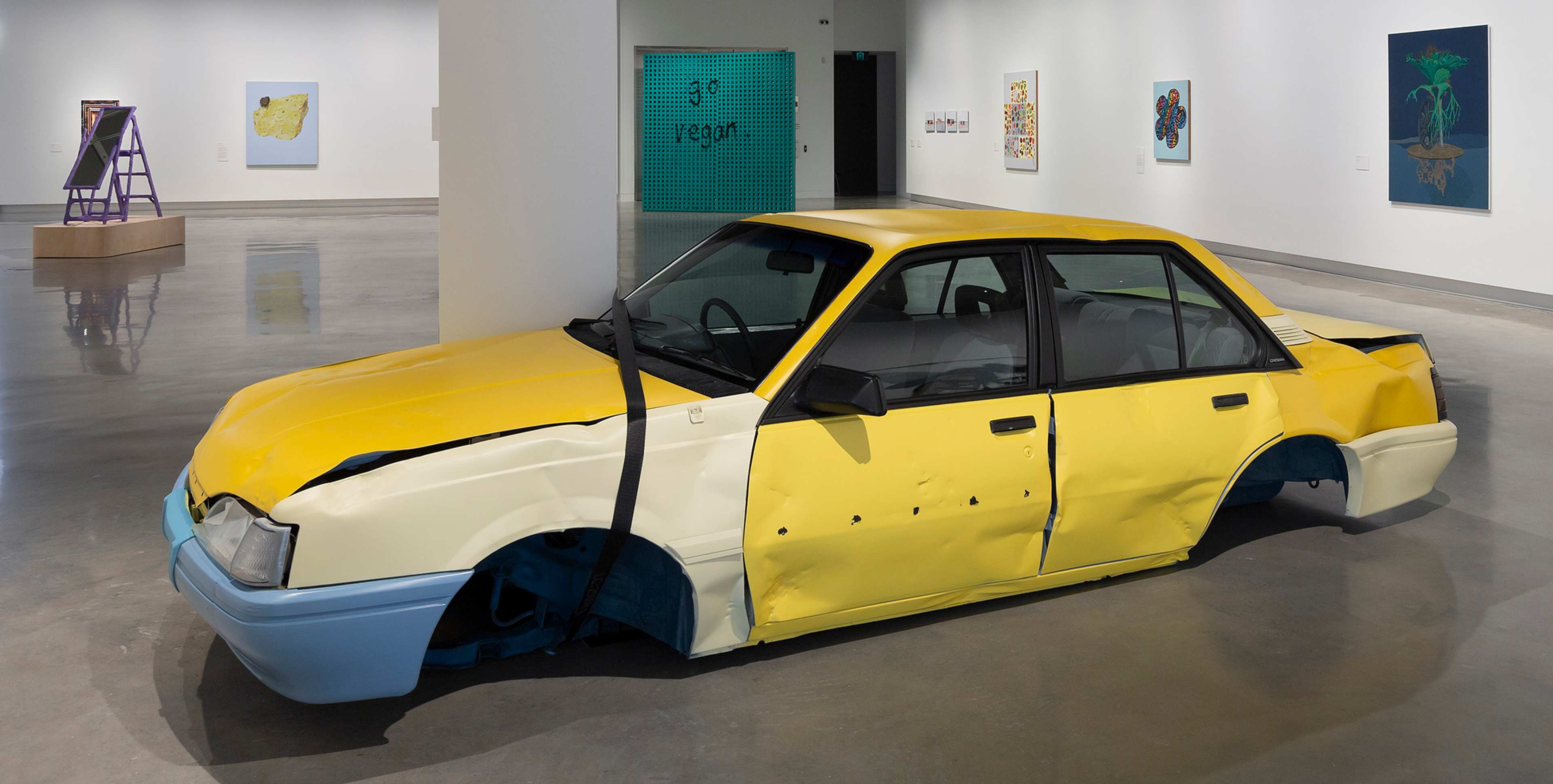 A yellow car with blue bumpers, heavily damaged and flattened, is displayed as an art installation in a modern gallery. The gallery has white walls and a polished concrete floor.