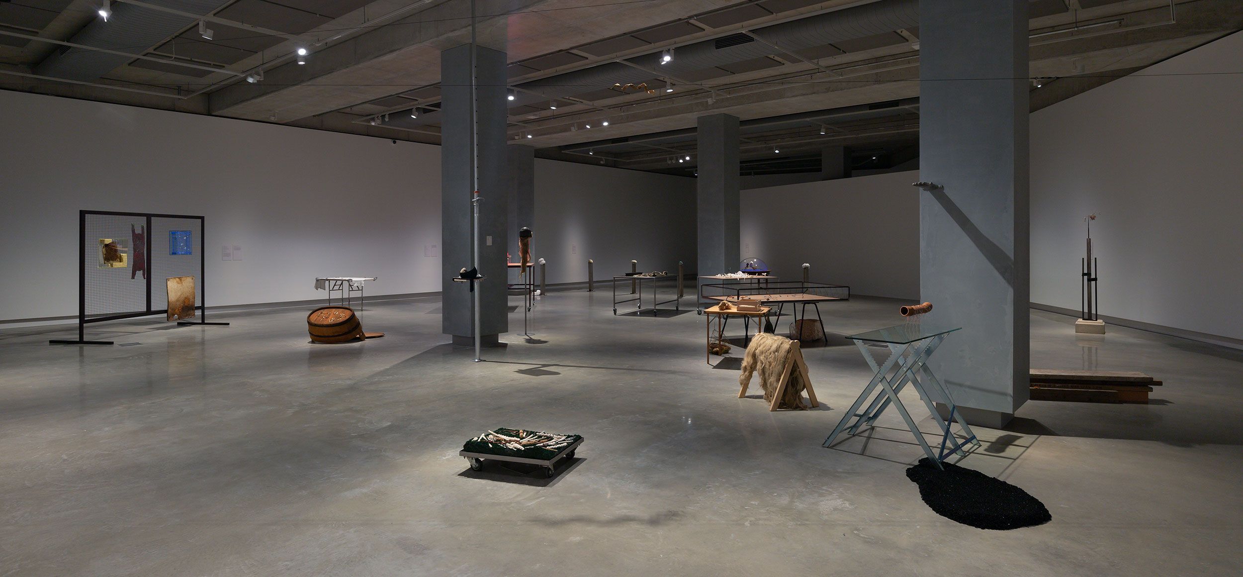 A modern art gallery with concrete floors and pillars displays various abstract sculptures and mixed-media installations spaced throughout the large, well-lit room.