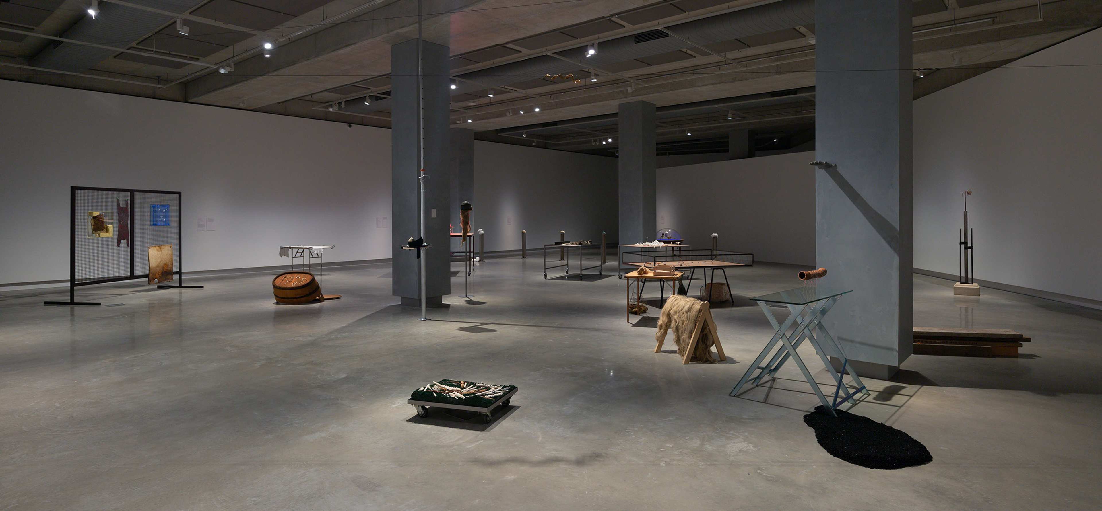 A modern art gallery with concrete floors and pillars displays various abstract sculptures and mixed-media installations spaced throughout the large, well-lit room.