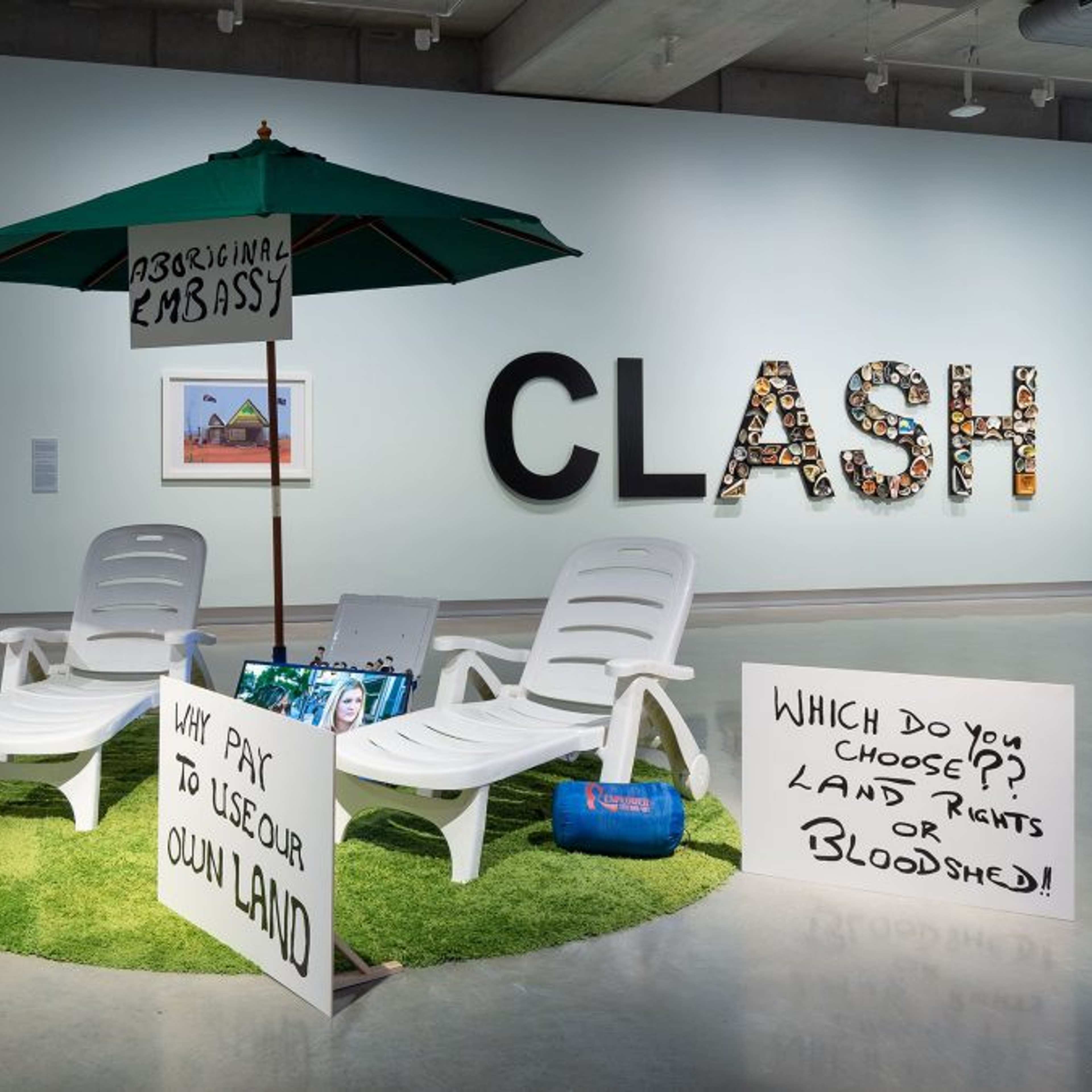 Art installation featuring white lounge chairs, an umbrella labeled Aboriginal Embassy, and protest signs on artificial grass. The signs discuss land rights. Framed photos and CLASH text are seen on the gallery wall behind.