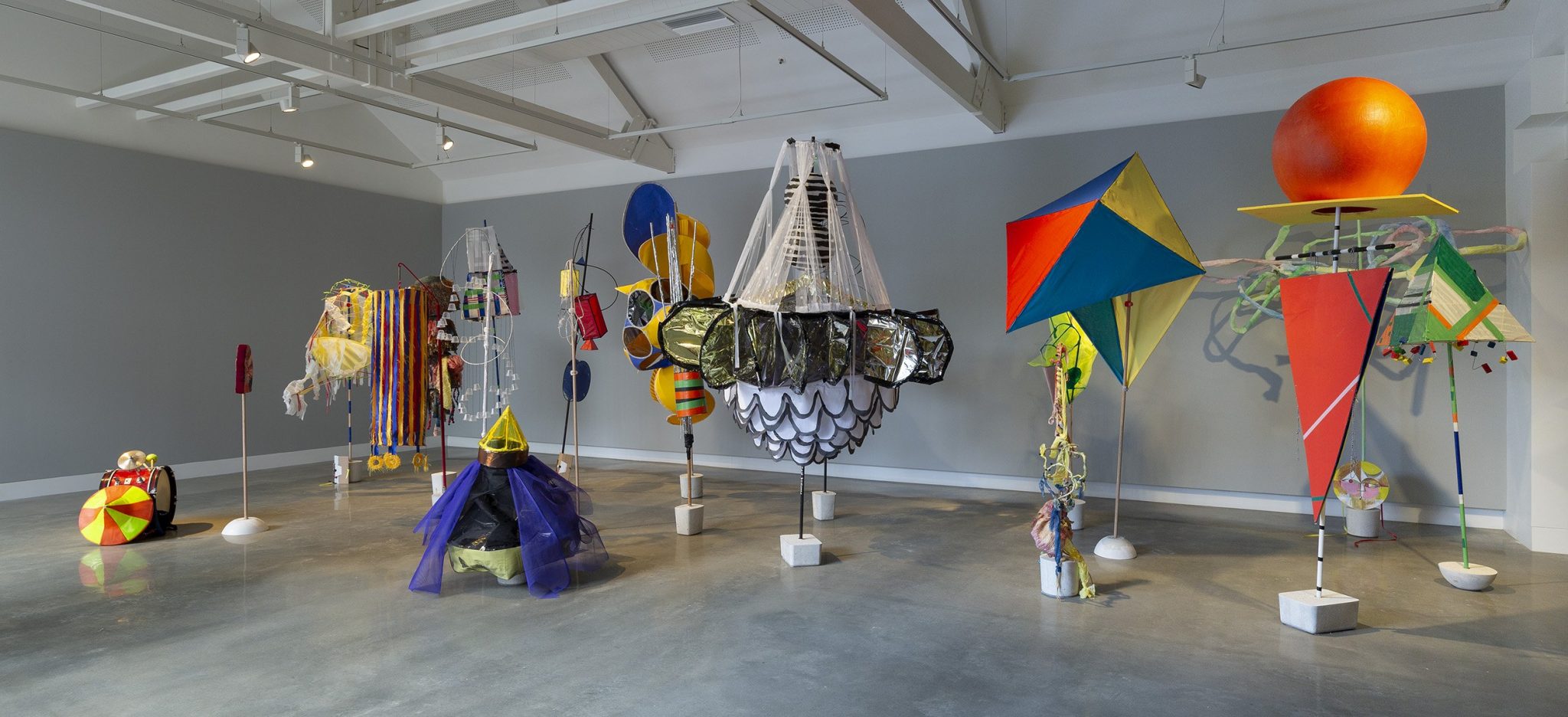 Colorful, abstract sculptures made from mixed materials including fabric, plastic, and metal are arranged in a well-lit gallery space with gray walls and a concrete floor. The sculptures vary in shapes and sizes.