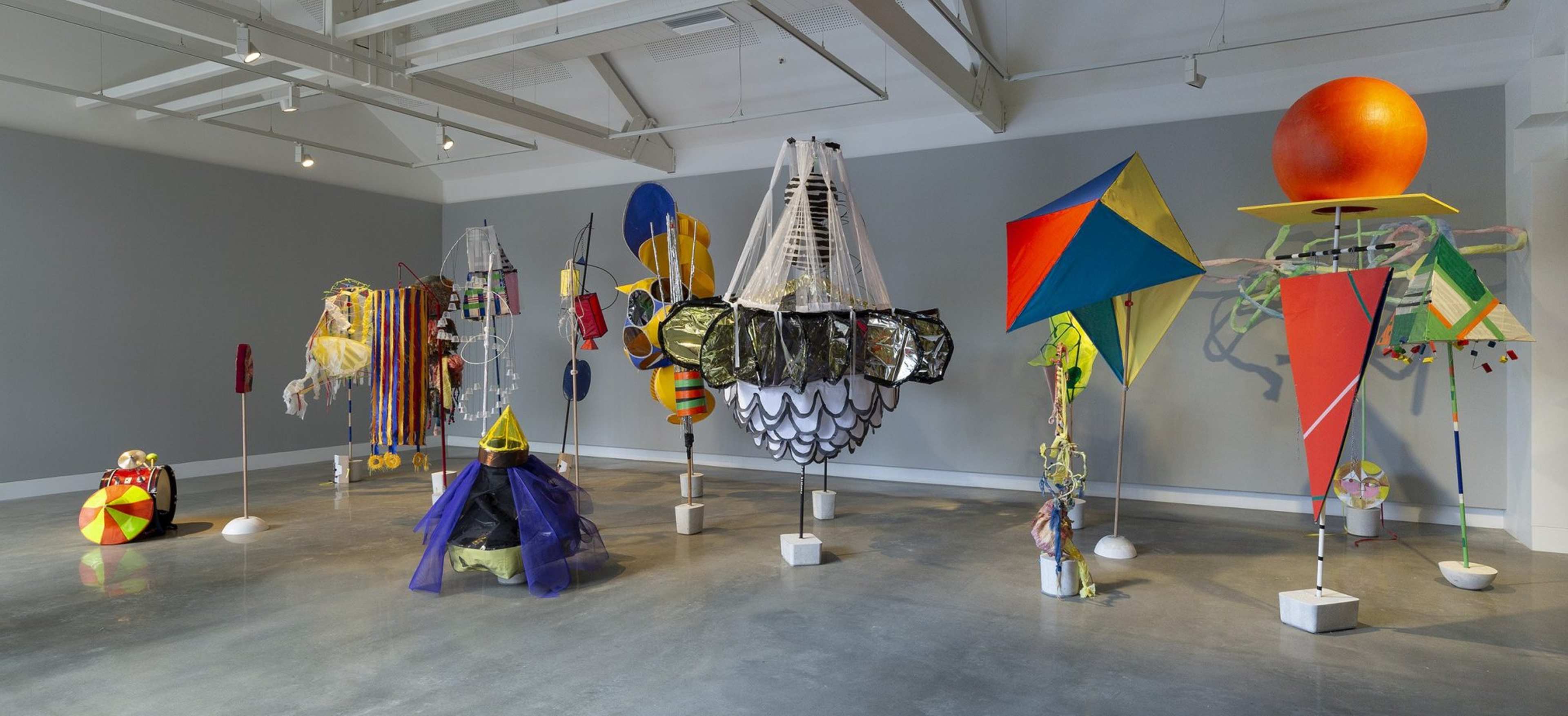 Colorful, abstract sculptures made from mixed materials including fabric, plastic, and metal are arranged in a well-lit gallery space with gray walls and a concrete floor. The sculptures vary in shapes and sizes.