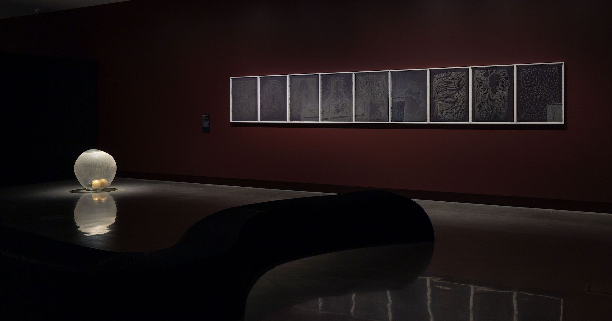 A dimly lit gallery with a glowing, round light sculpture on the floor and a series of framed, monochromatic artworks displayed on a dark red wall. The polished floor and curved bench enhance the reflective atmosphere.