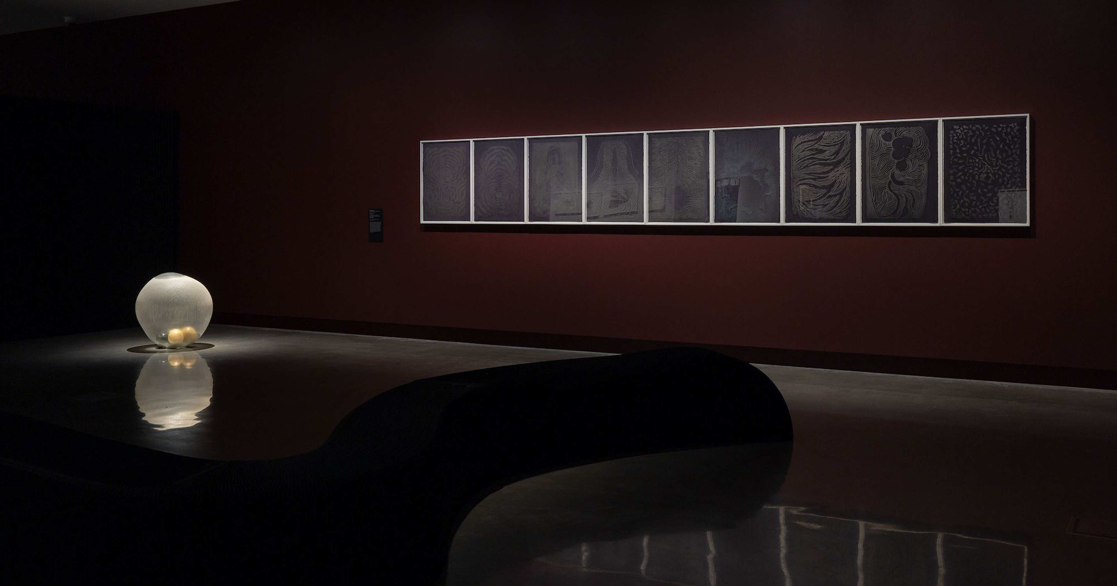 A dimly lit gallery with a glowing, round light sculpture on the floor and a series of framed, monochromatic artworks displayed on a dark red wall. The polished floor and curved bench enhance the reflective atmosphere.