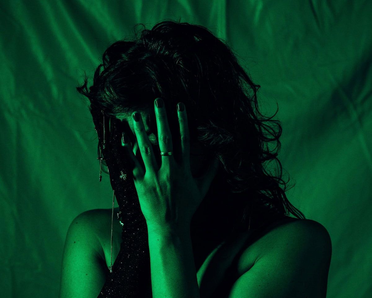 A person under green lighting, with long dark curly hair, covering their face with their hands, wearing on black, sparkly glove.